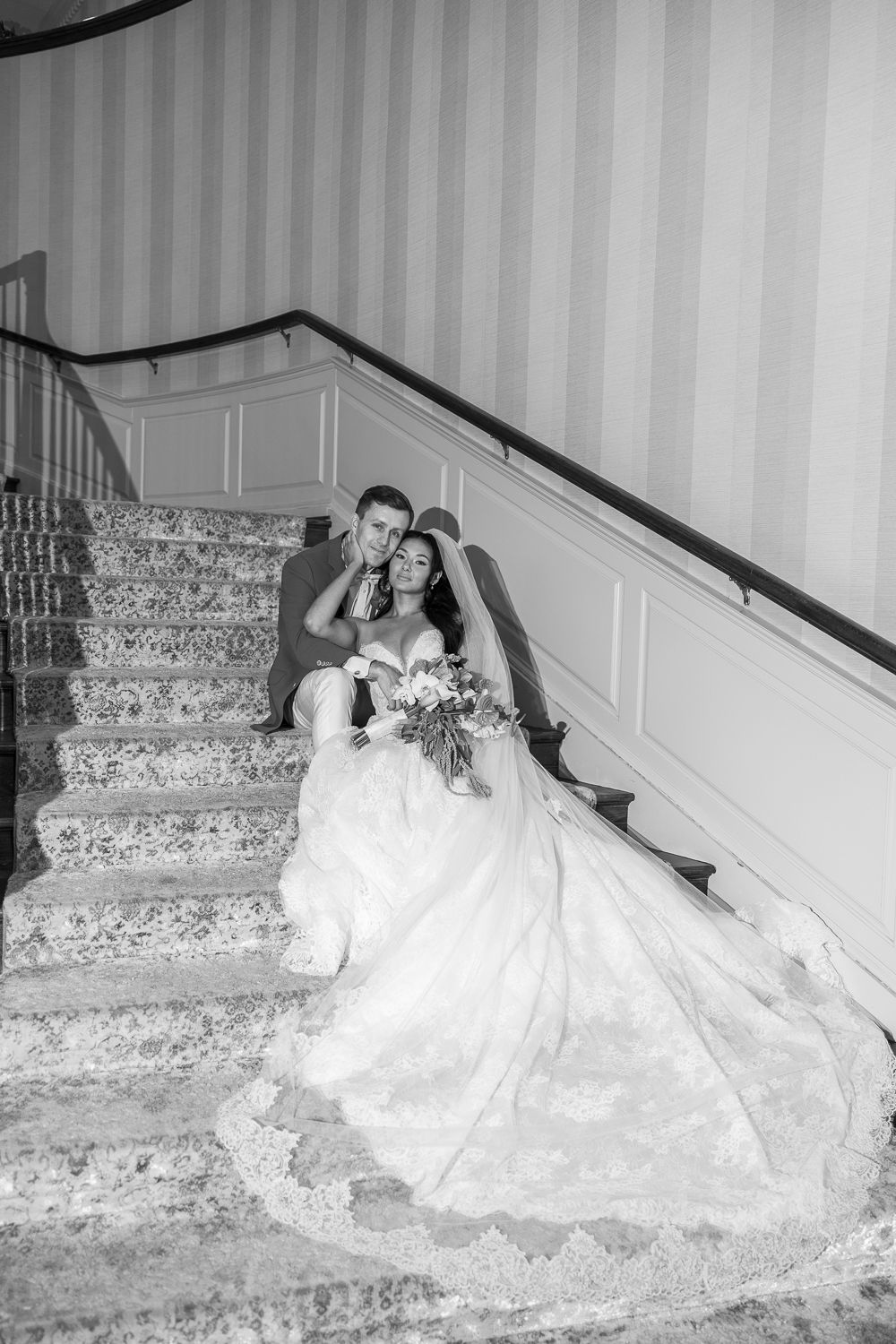Black and white photo of bride and groom sitting on elegant staircase, bride in lace gown with bouquet.