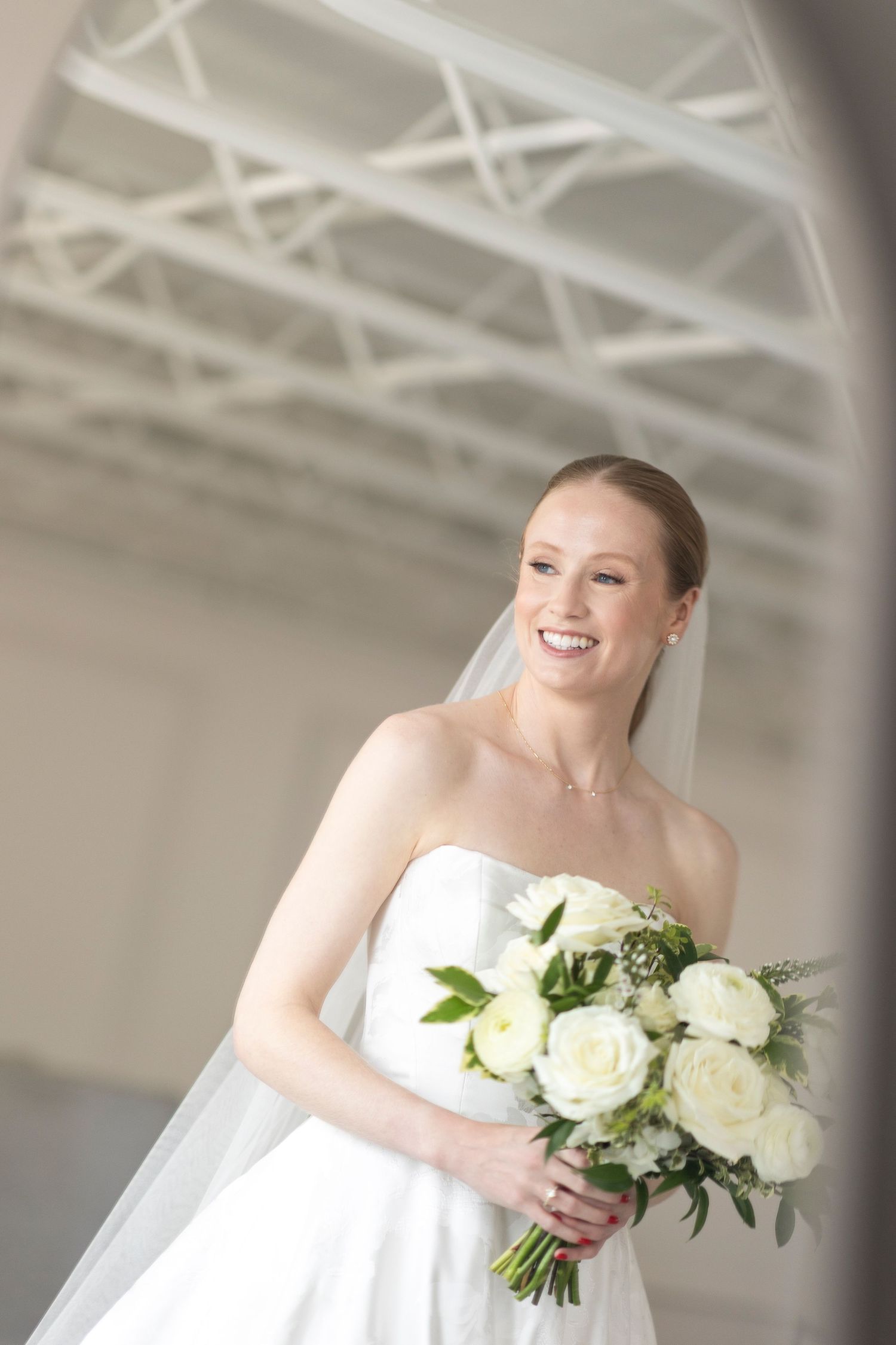 A bride in a white strapless gown holds a bouquet of white roses and greenery against an industrial white backdrop.