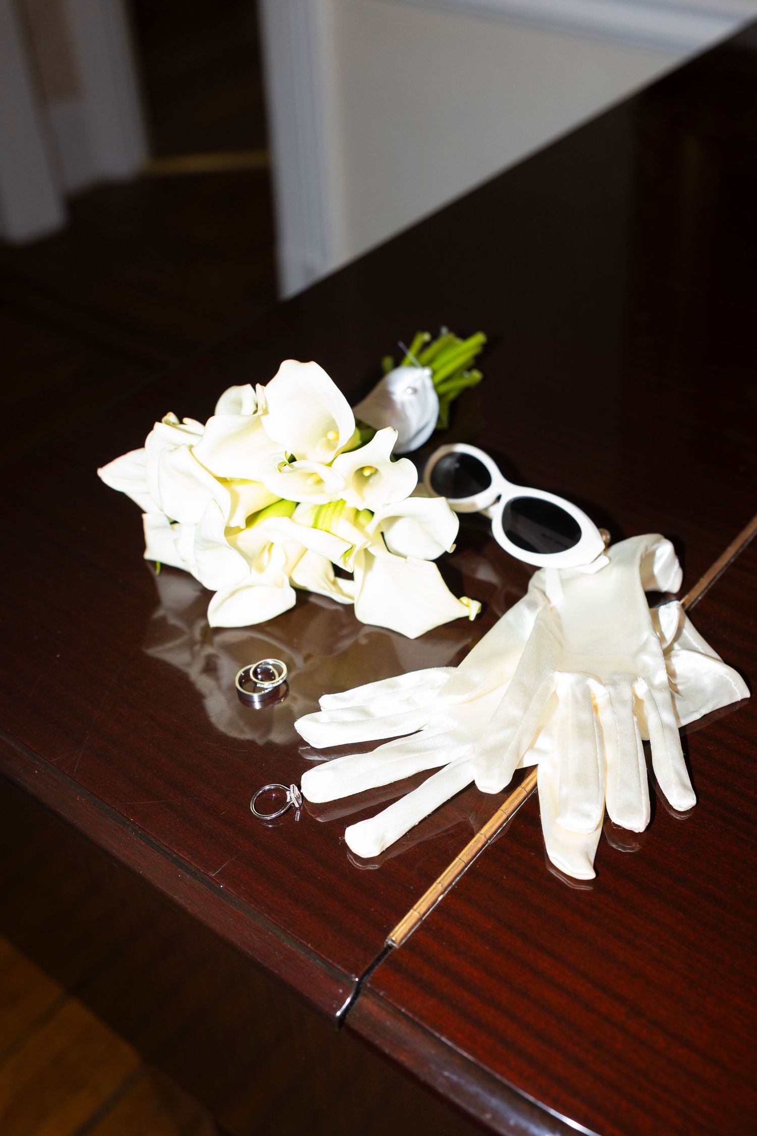 Wedding accessories including white gloves, flowers and rings arranged on wooden surface.