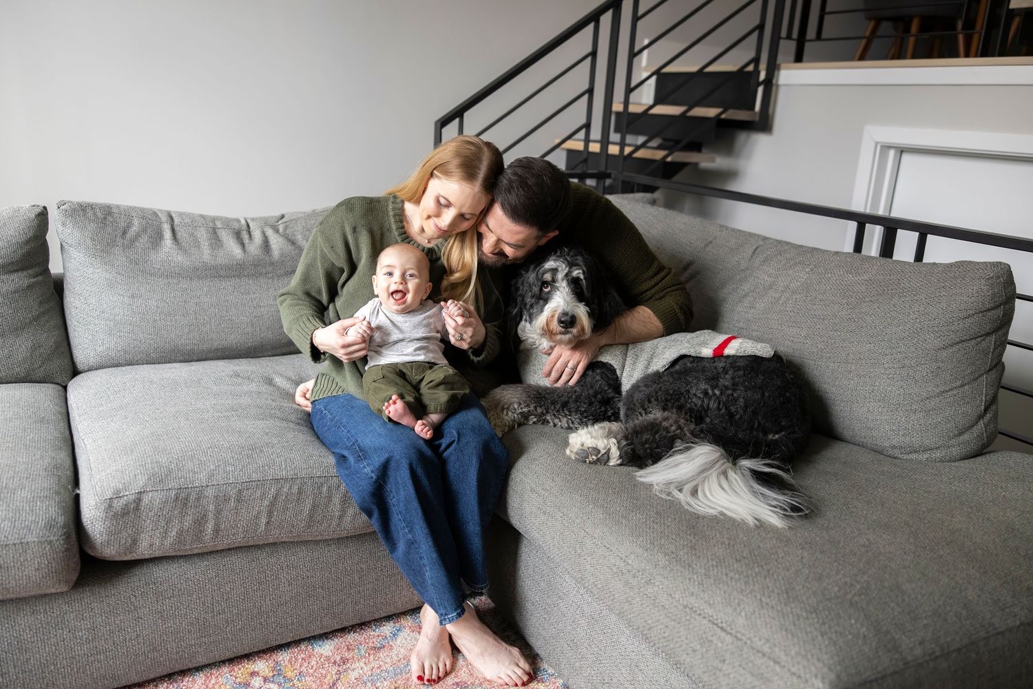 A couple relaxes on a grey couch with their dog in a modern living room setting.