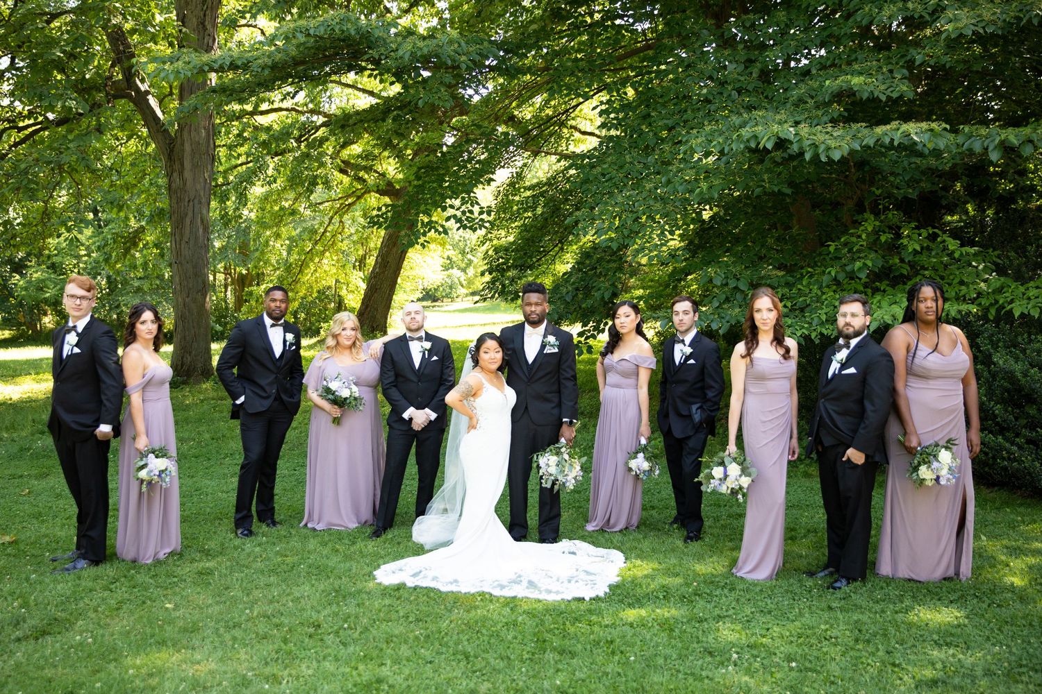 Wedding party poses for formal photos on grass under leafy green trees.