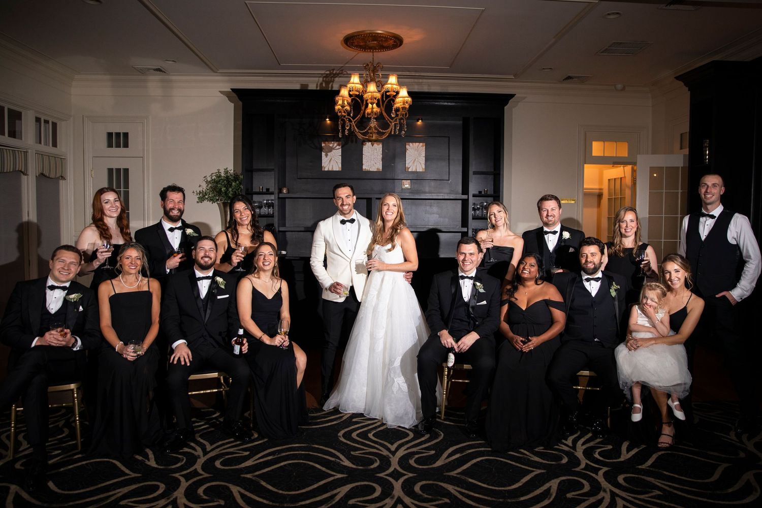 Wedding party in black attire poses with bride and groom in elegant venue with chandelier and fireplace.