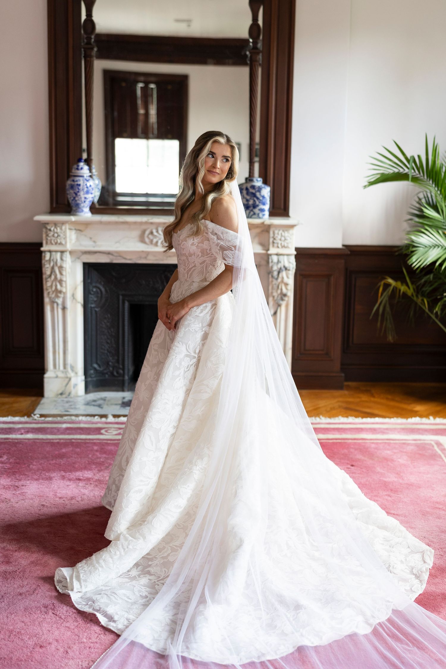Bride in a flowing lace wedding gown poses on pink carpet near an elegant fireplace.