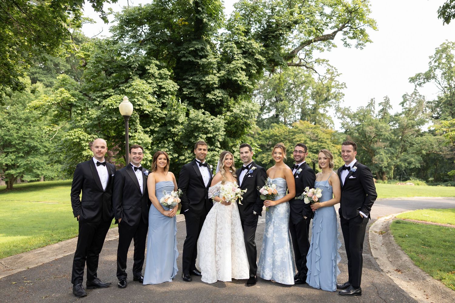 Wedding party in formal attire posing outdoors under green trees on a sunny day.