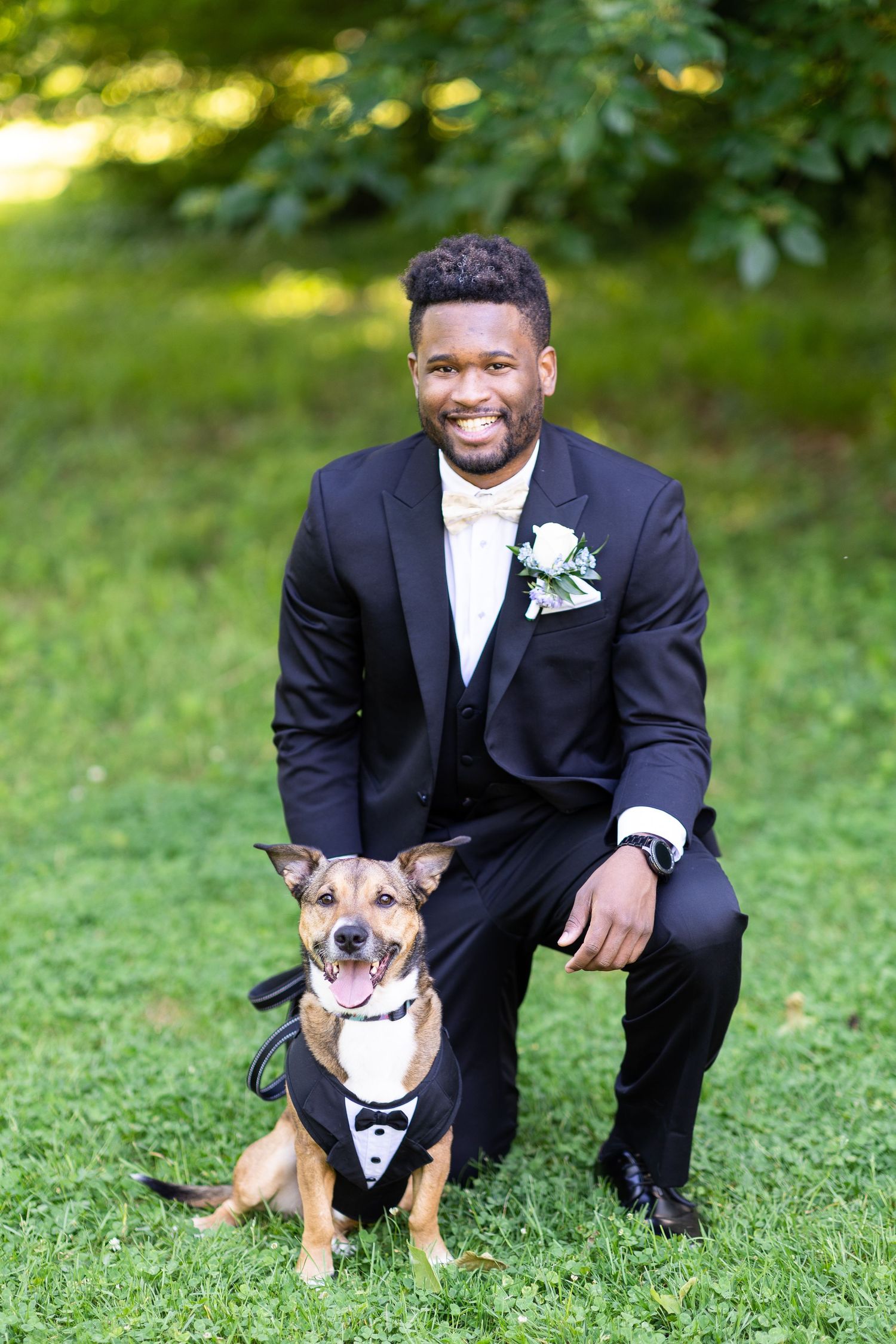 Groomsman in black tuxedo poses with small brown dog wearing bowtie on grass.