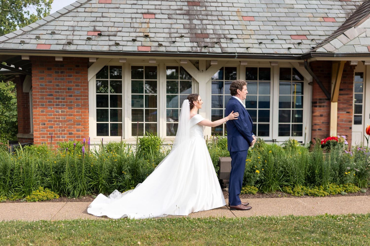 A couple in wedding attire share a romantic moment outside a brick building with a shingled roof.