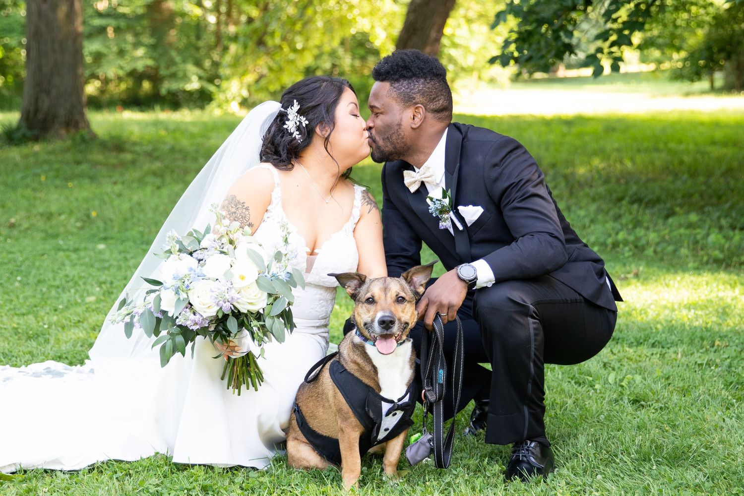 Wedding couple poses with their pet dog on a grassy lawn during summer.