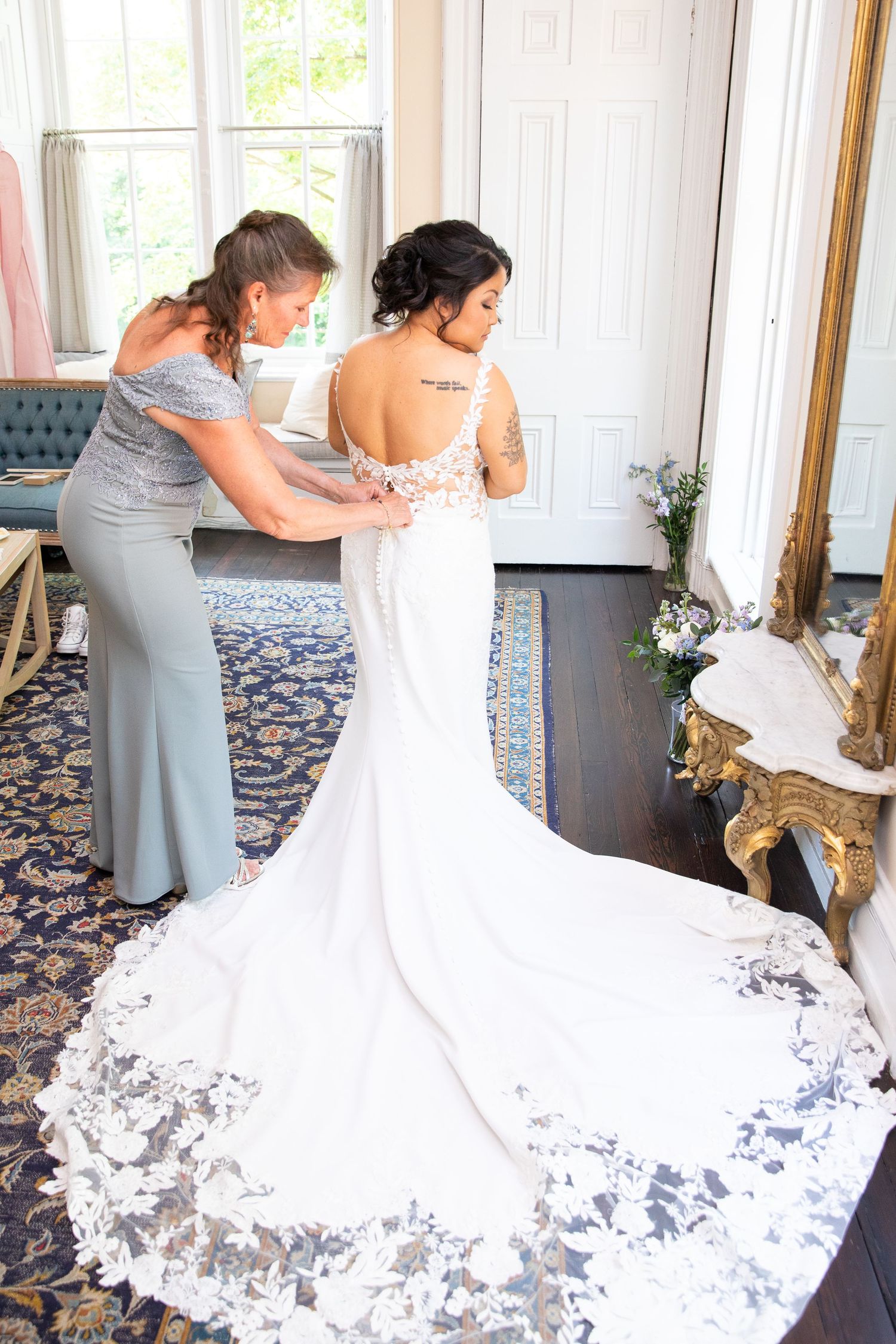 A bride in a white lace wedding gown with a flowing train gets help with final dress adjustments.