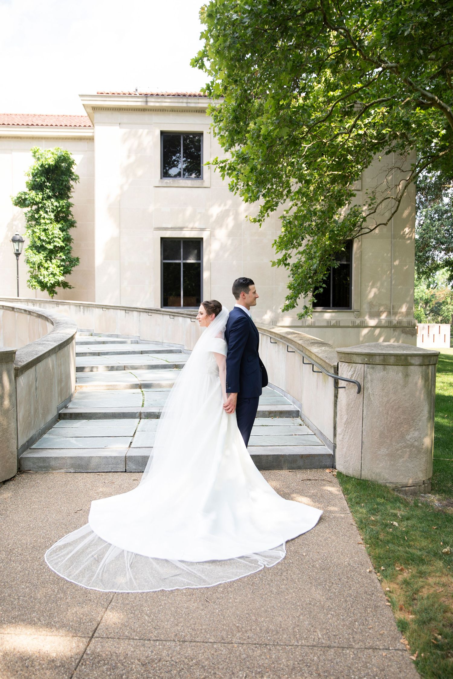 A wedding couple stands on stone steps in front of a classic white building with a flowing cathedral veil.