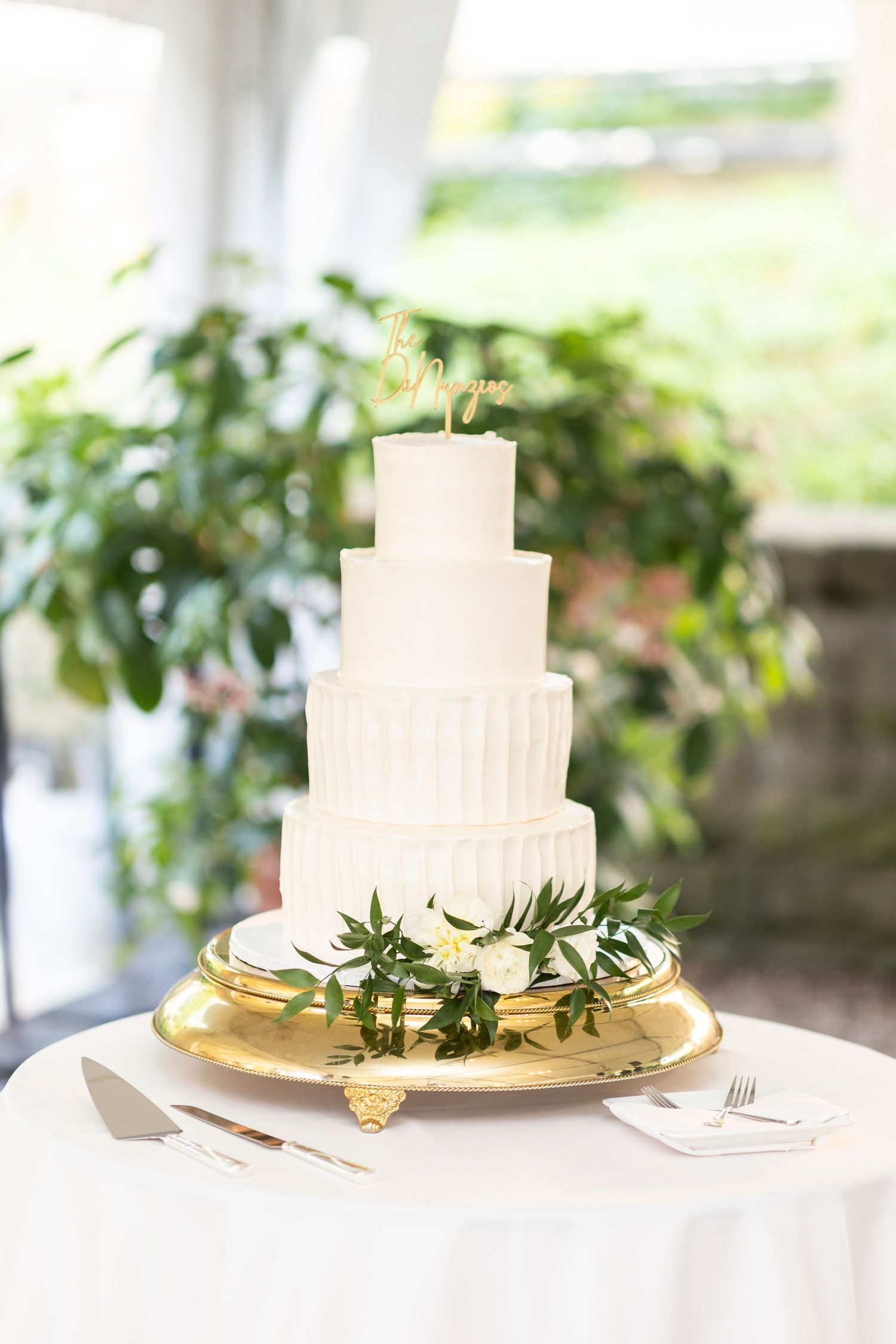 Elegant white tiered wedding cake decorated with greenery on a gold cake stand.