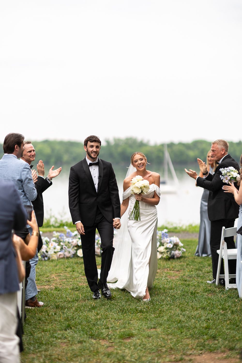 Bride and groom walk down outdoor aisle holding hands, guests applaud, waterfront backdrop with sailboat.
