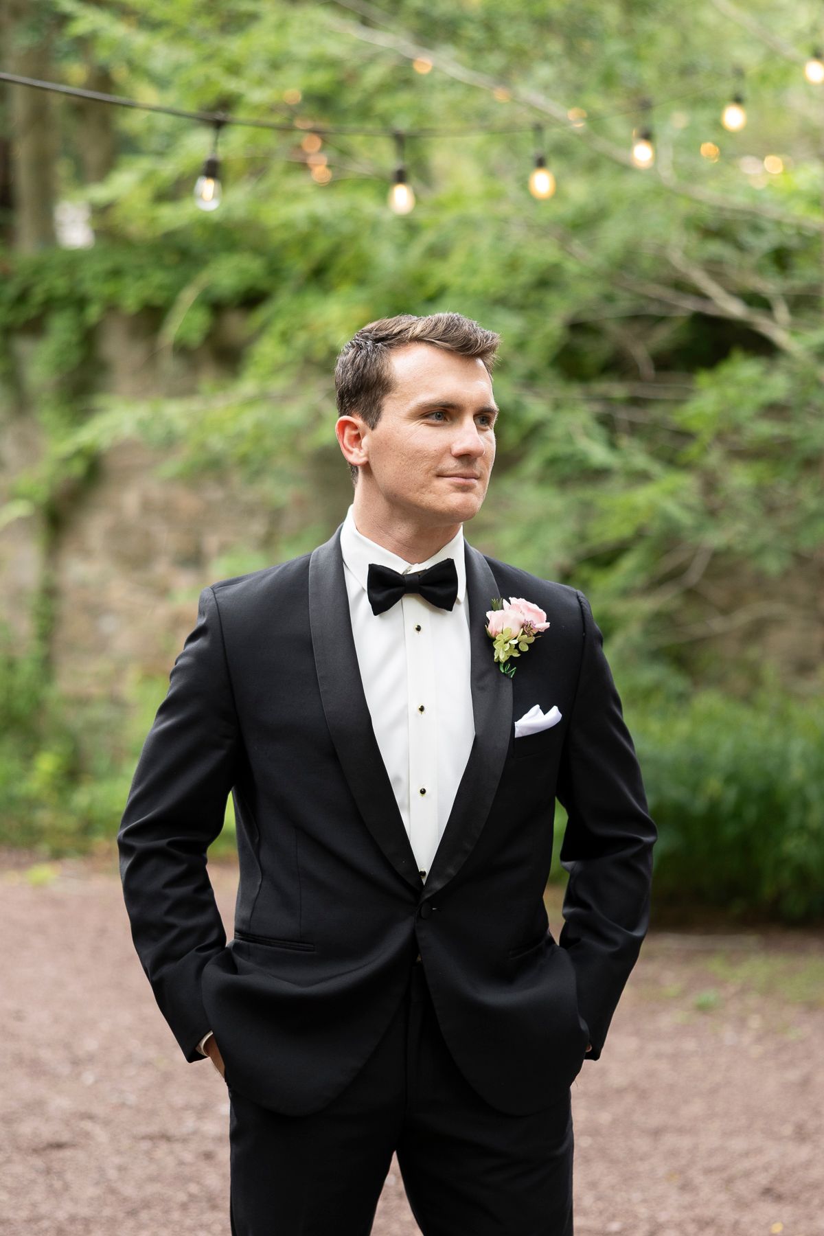 Groom in black tuxedo with bow tie and boutonniere stands outdoors with hands in pockets at wedding.