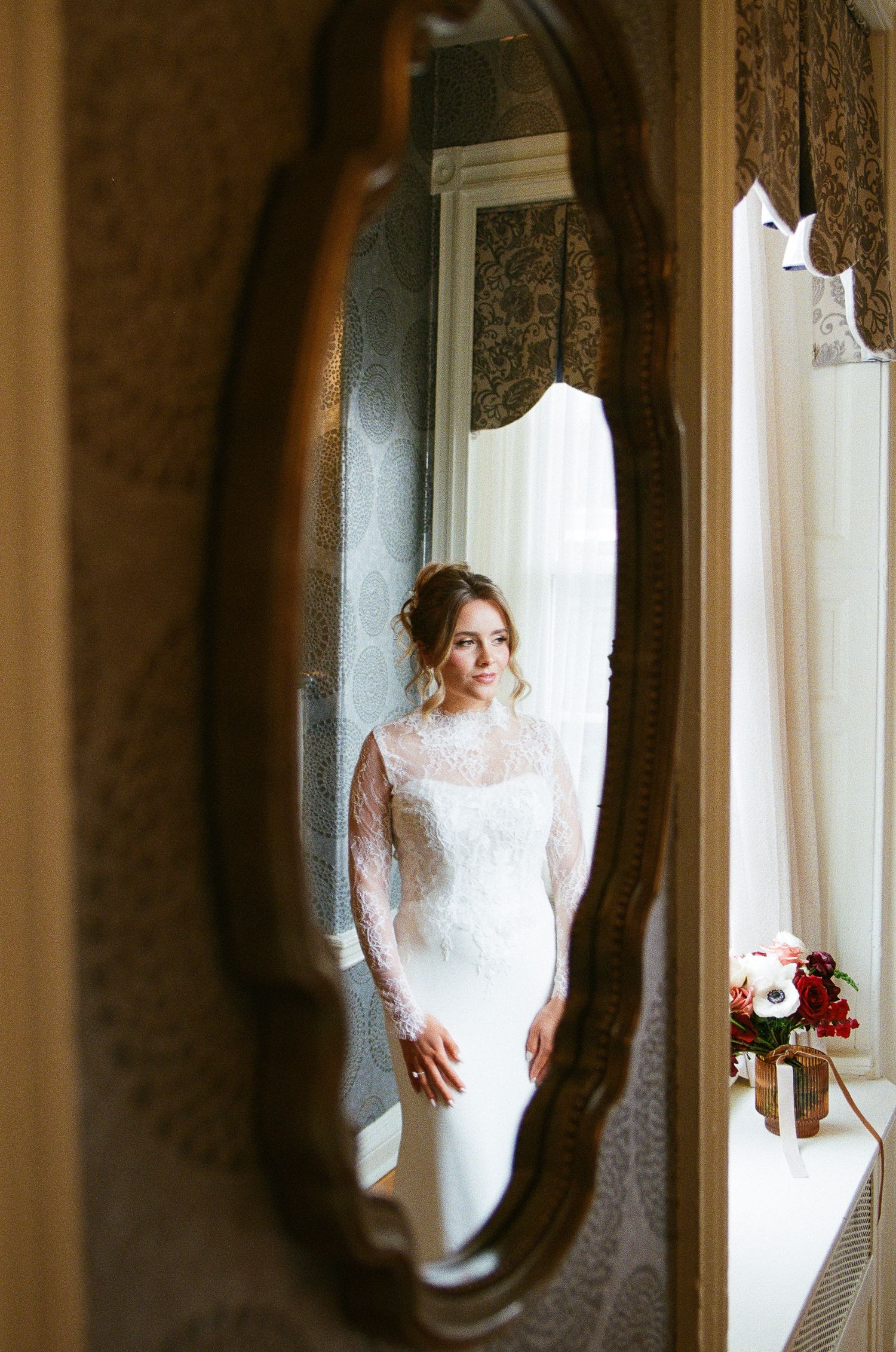 Bride in lace long-sleeve wedding gown reflected in ornate vintage mirror, with floral bouquet on windowsill.