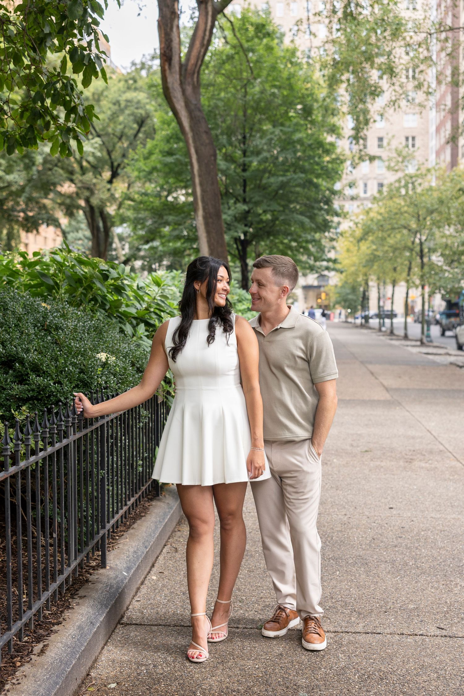 A couple strolls along a tree-lined pathway wearing coordinated beige and white outfits.