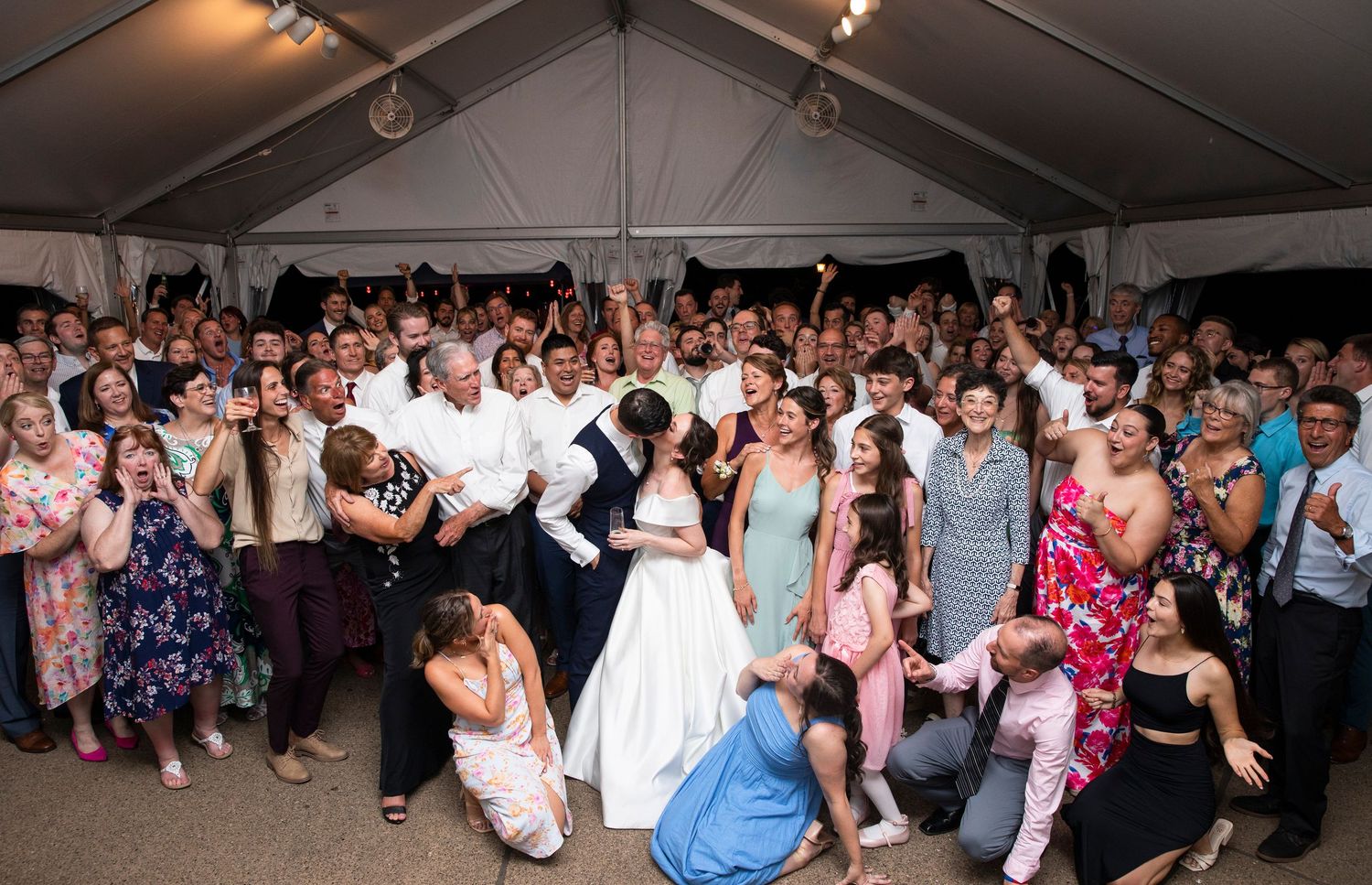 Large group of wedding guests dancing together under a white tent at night.