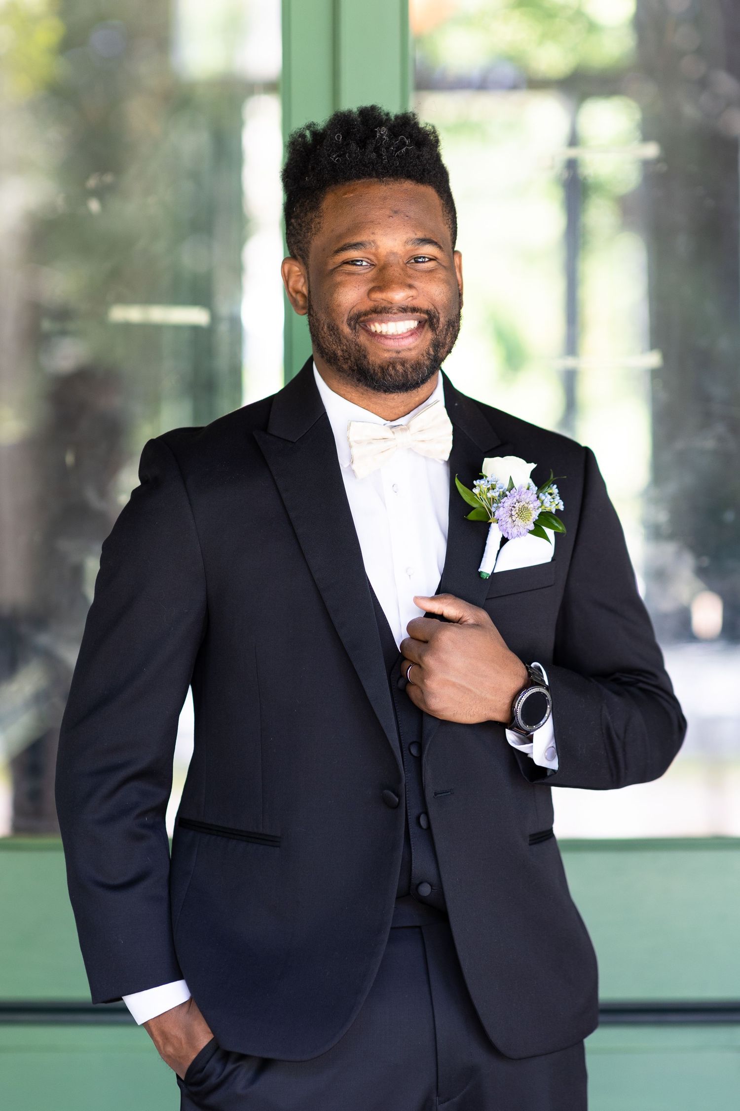 A groom in a classic black tuxedo with purple boutonniere poses against a green background.