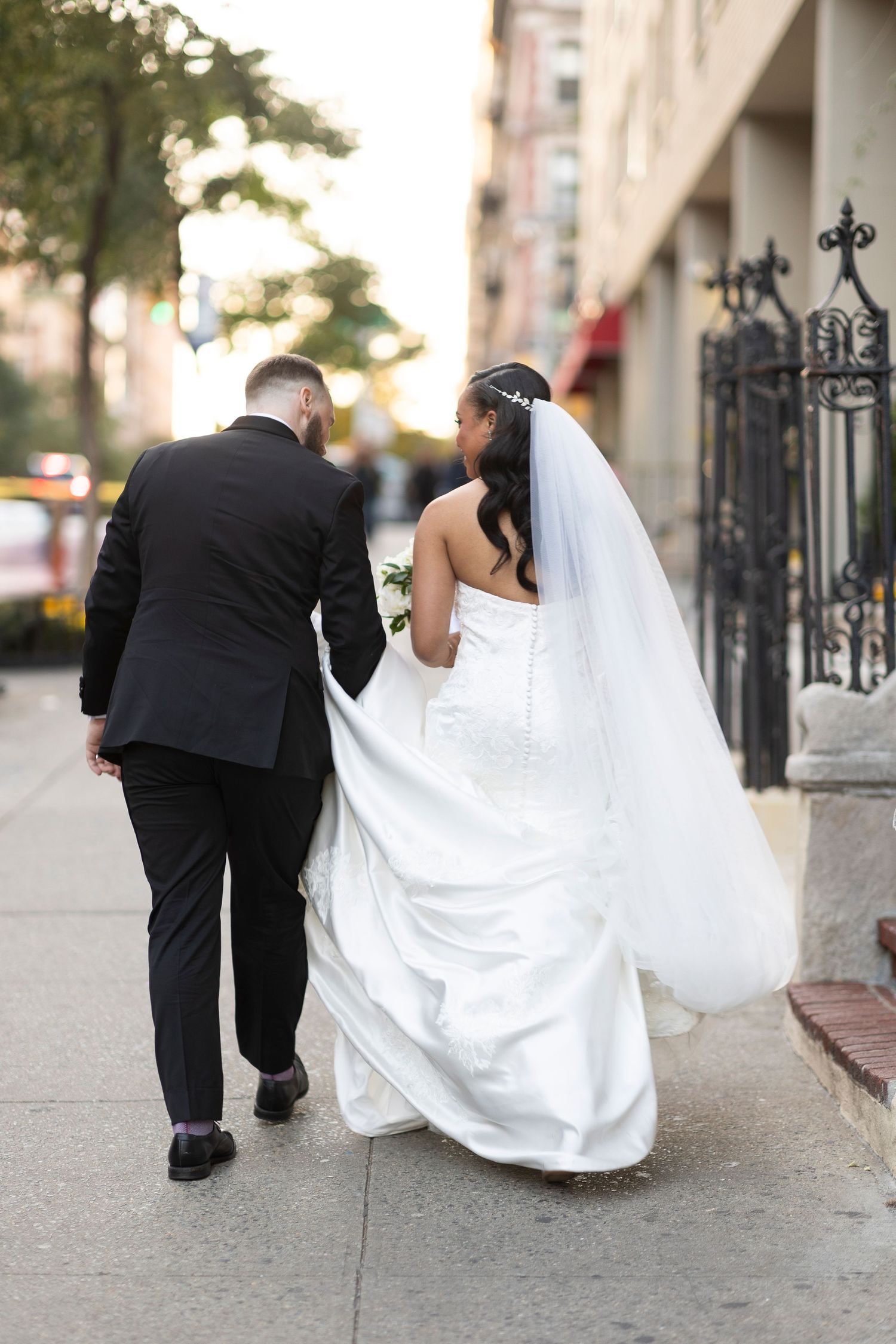 Newlyweds walking down a city sidewalk with bride's flowing white dress train trailing behind.
