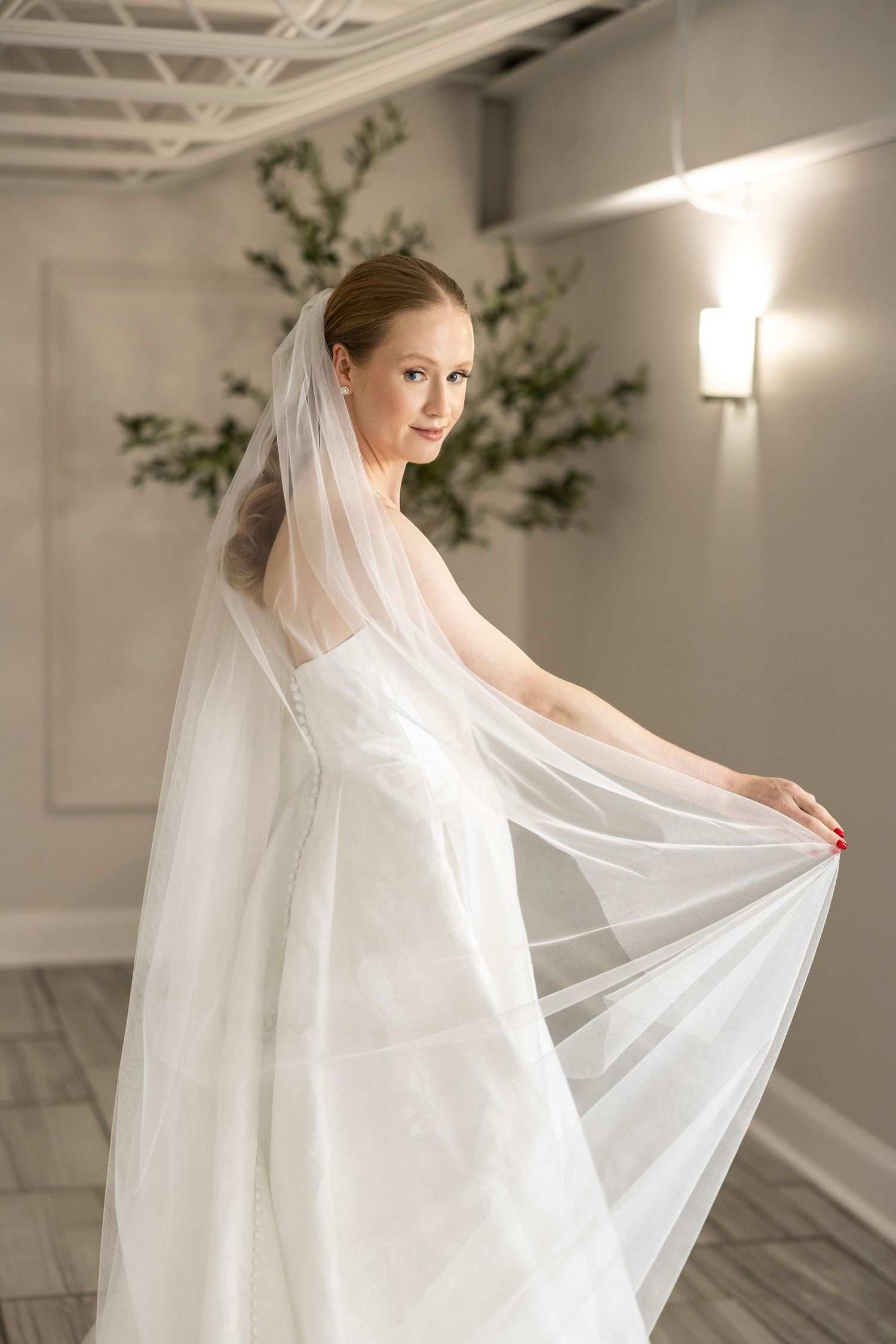 Bride wearing flowing veil poses gracefully in white wedding gown against neutral studio backdrop.