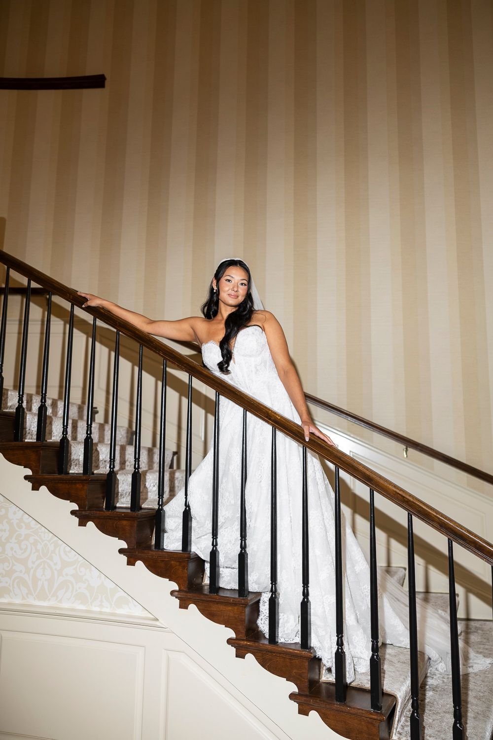 Elegant bride in white lace gown and veil poses on curved wooden staircase with black iron railing in luxurious venue.