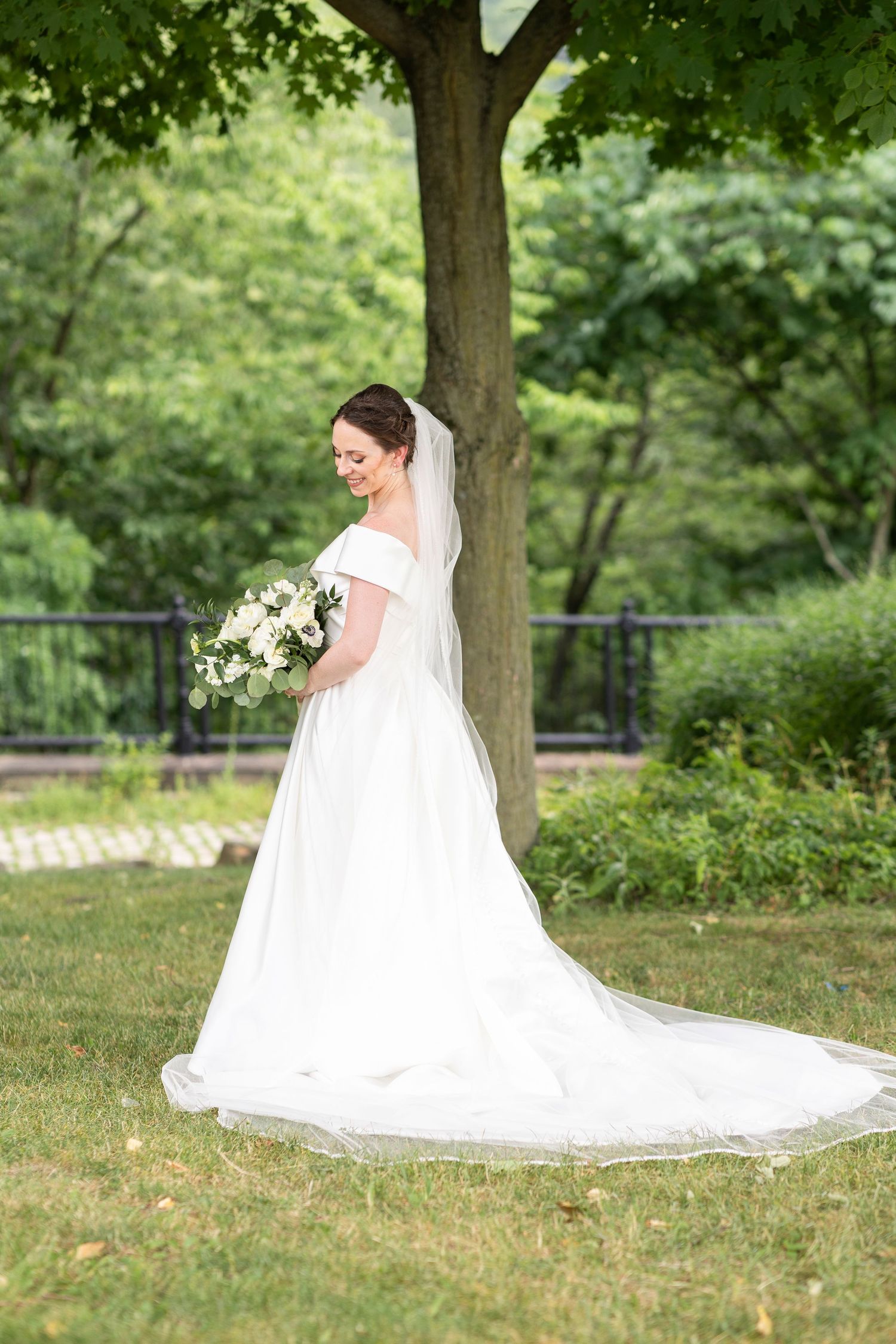 Bride in flowing white gown with long train poses under trees in garden setting.