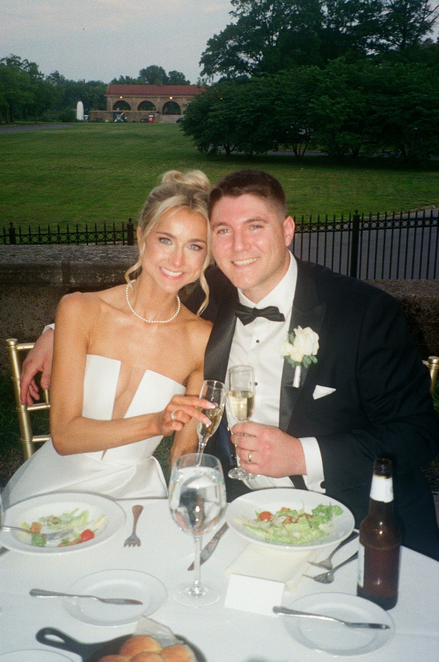 Bride and groom in formal attire toasting with champagne at outdoor wedding reception dinner table.