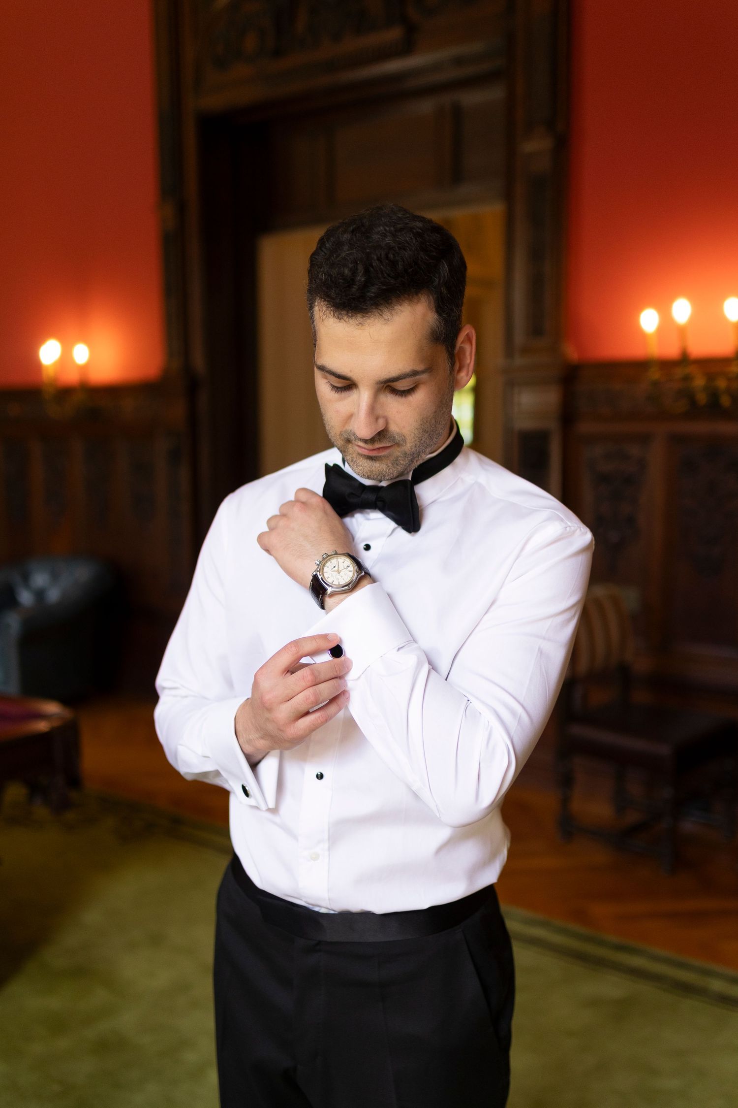 Man in formal attire adjusting white bowtie in warm lighting.