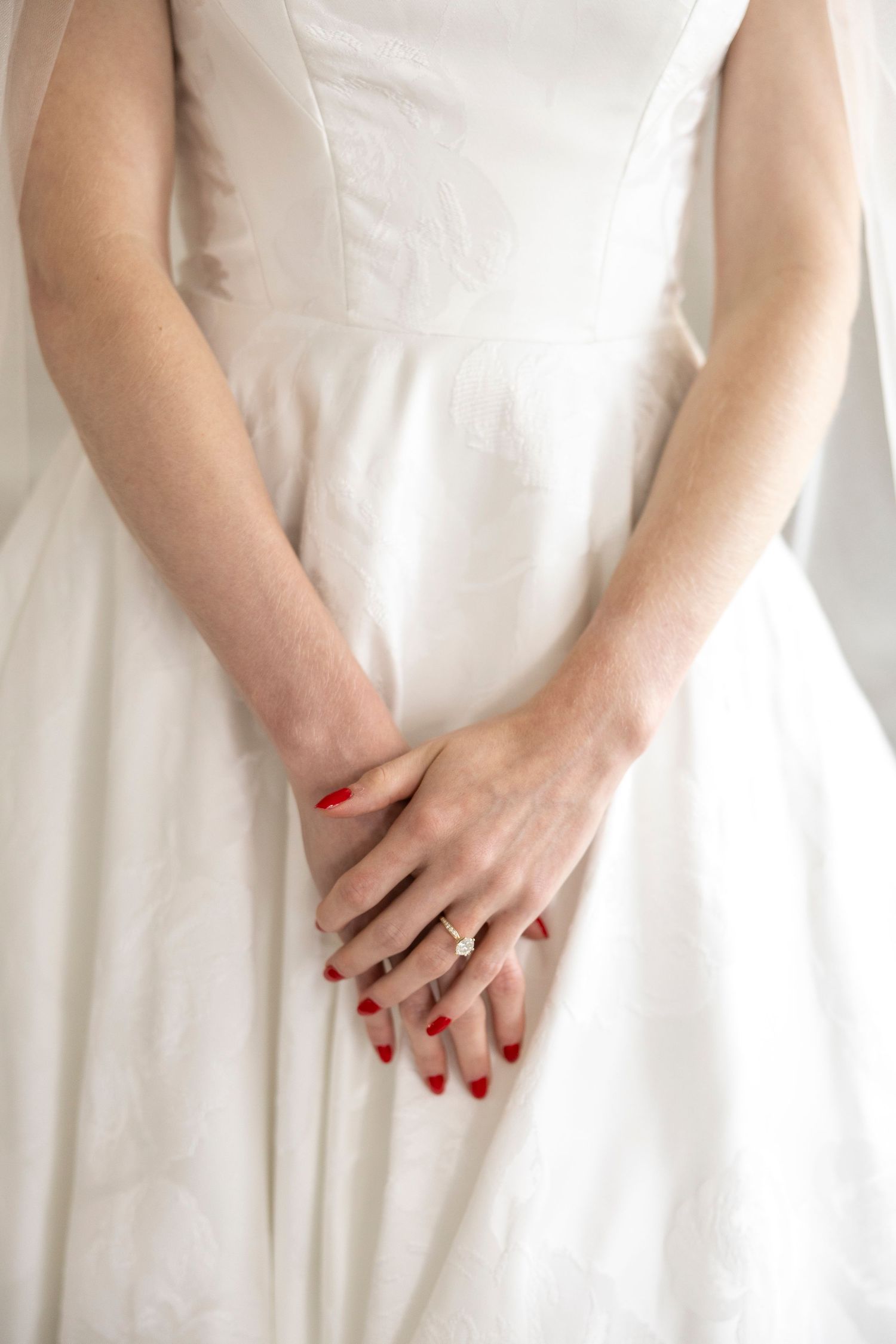 Close-up of hands with red manicure and wedding ring resting on white wedding dress.