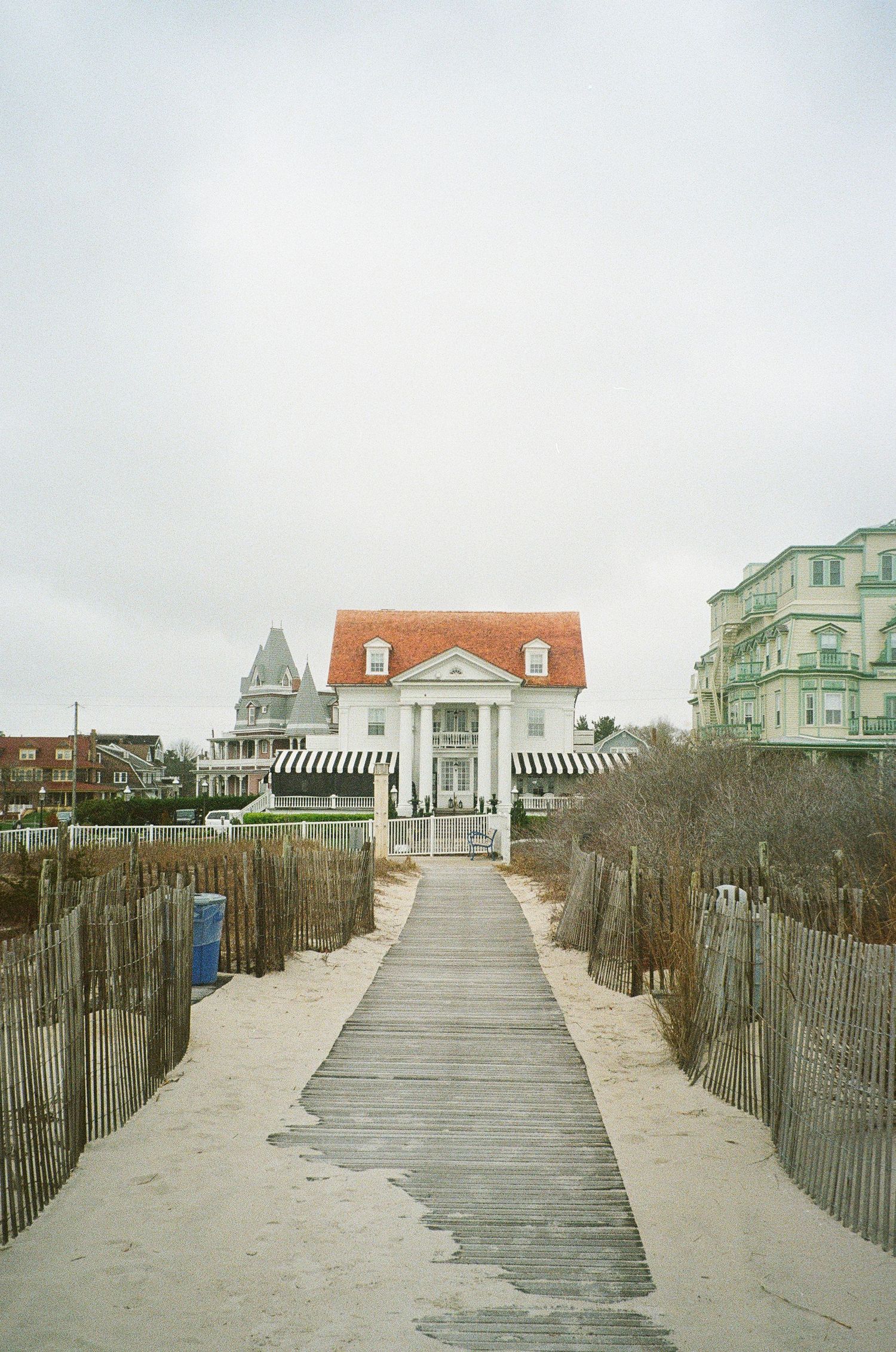 Wooden boardwalk path through beach dunes leading to coastal houses with orange roofs.