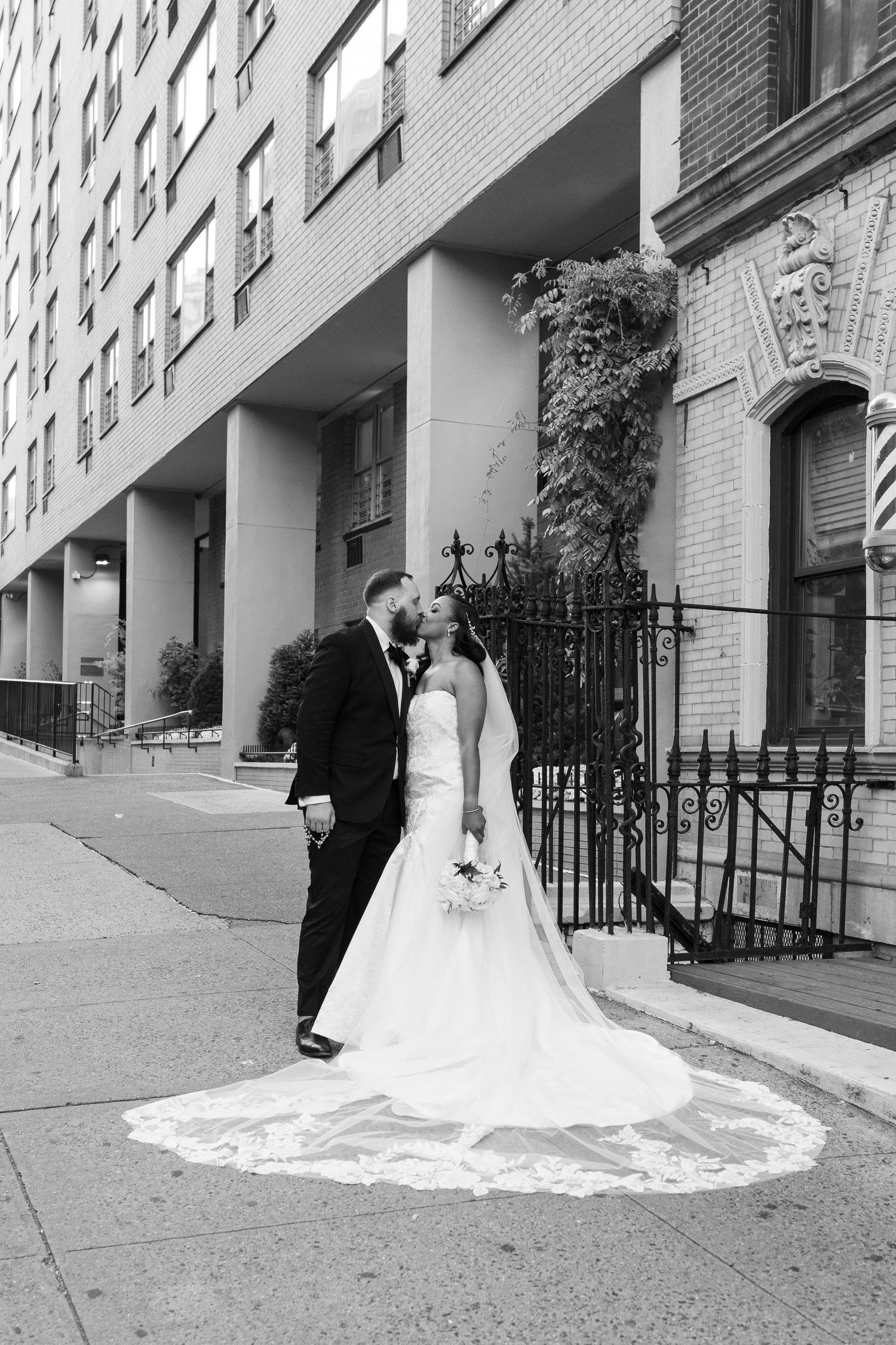 Black and white wedding photo with dramatic train against city building backdrop.