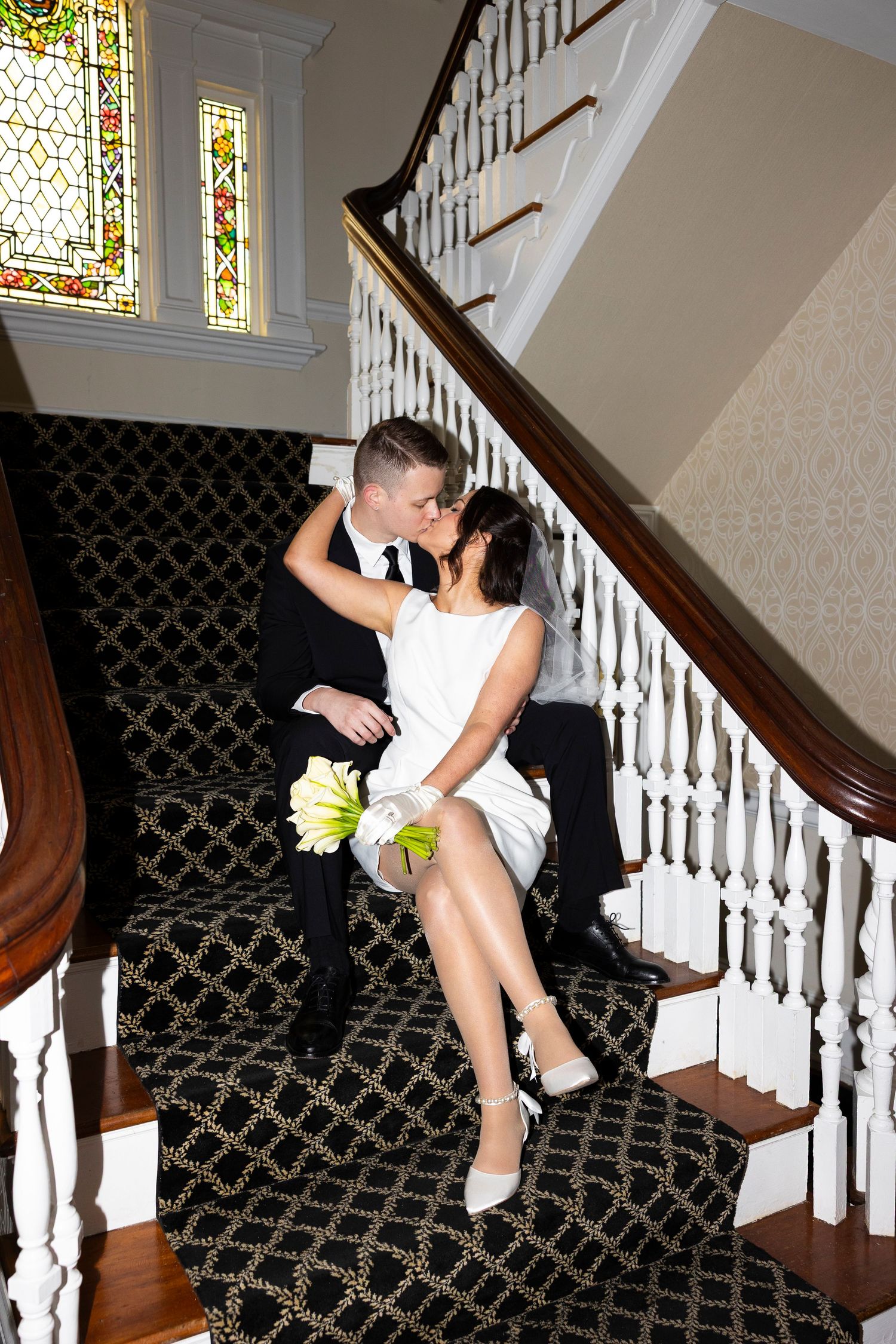 A couple shares a romantic moment on an elegant staircase with stained glass windows.