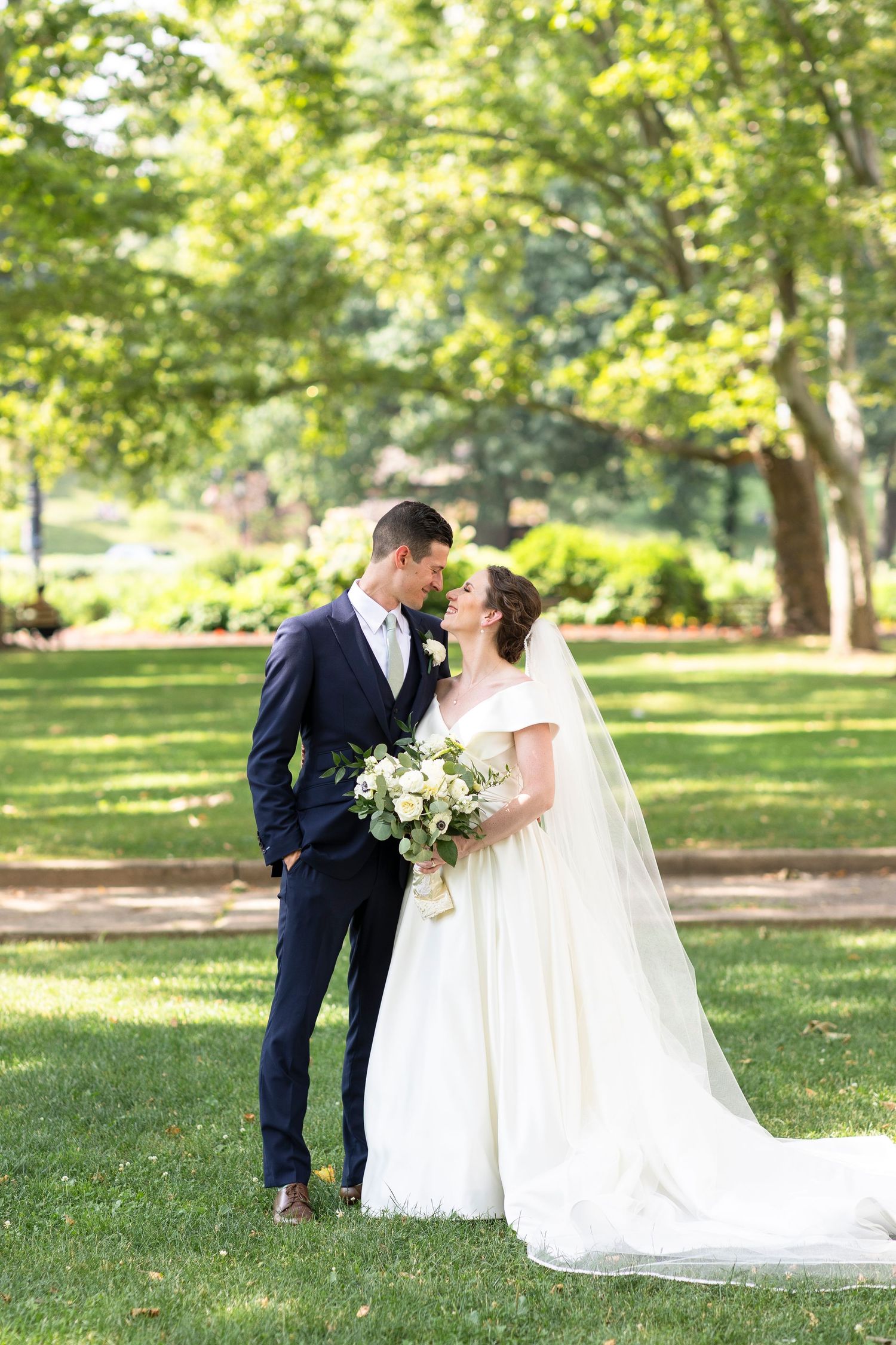 A romantic wedding couple shares intimate moments in a sunlit garden setting with flowing white dress and navy suit.