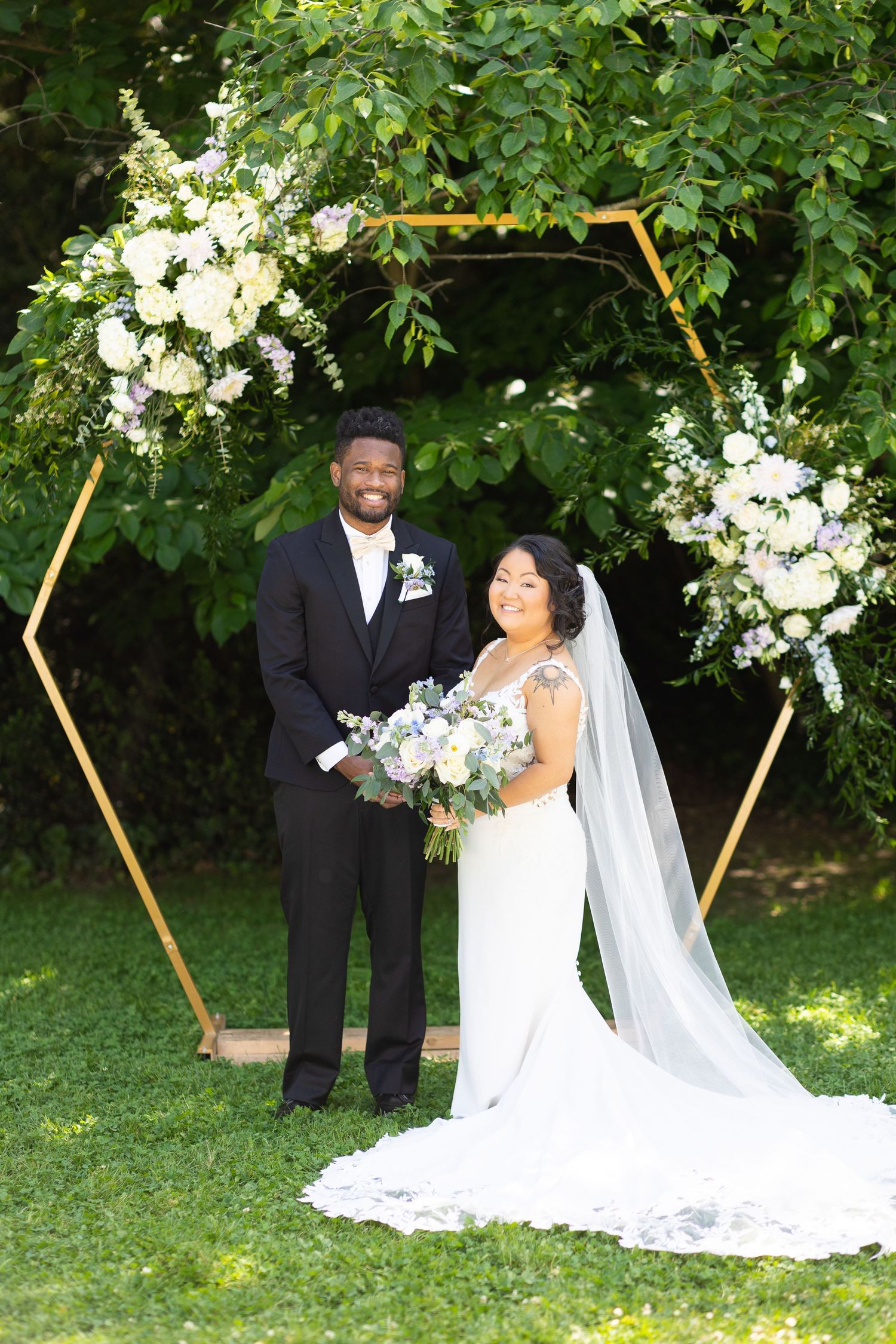 A couple poses under a hexagonal arch decorated with white flowers in a garden setting.