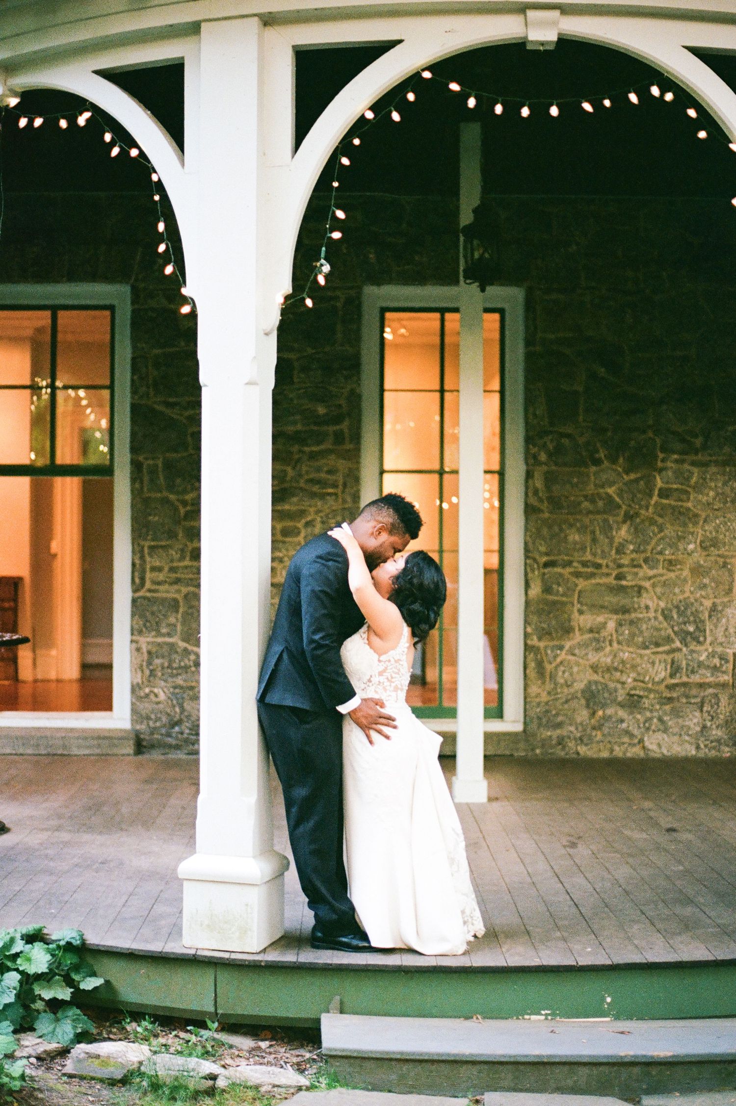 Newlyweds share intimate moments on wraparound porch with stone walls and white columns.