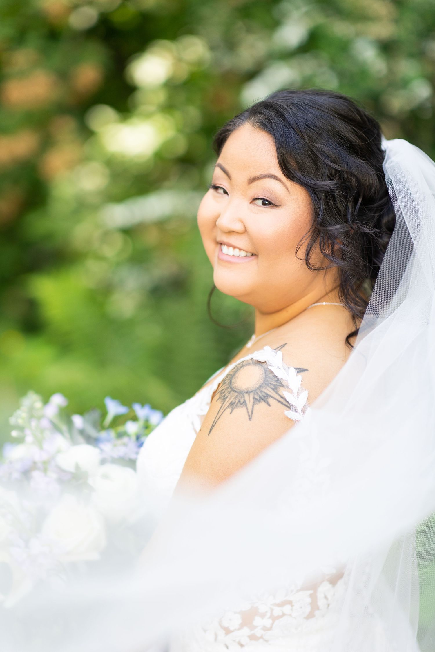 A bride wearing a white lace gown and veil smiles brightly against a green outdoor background.
