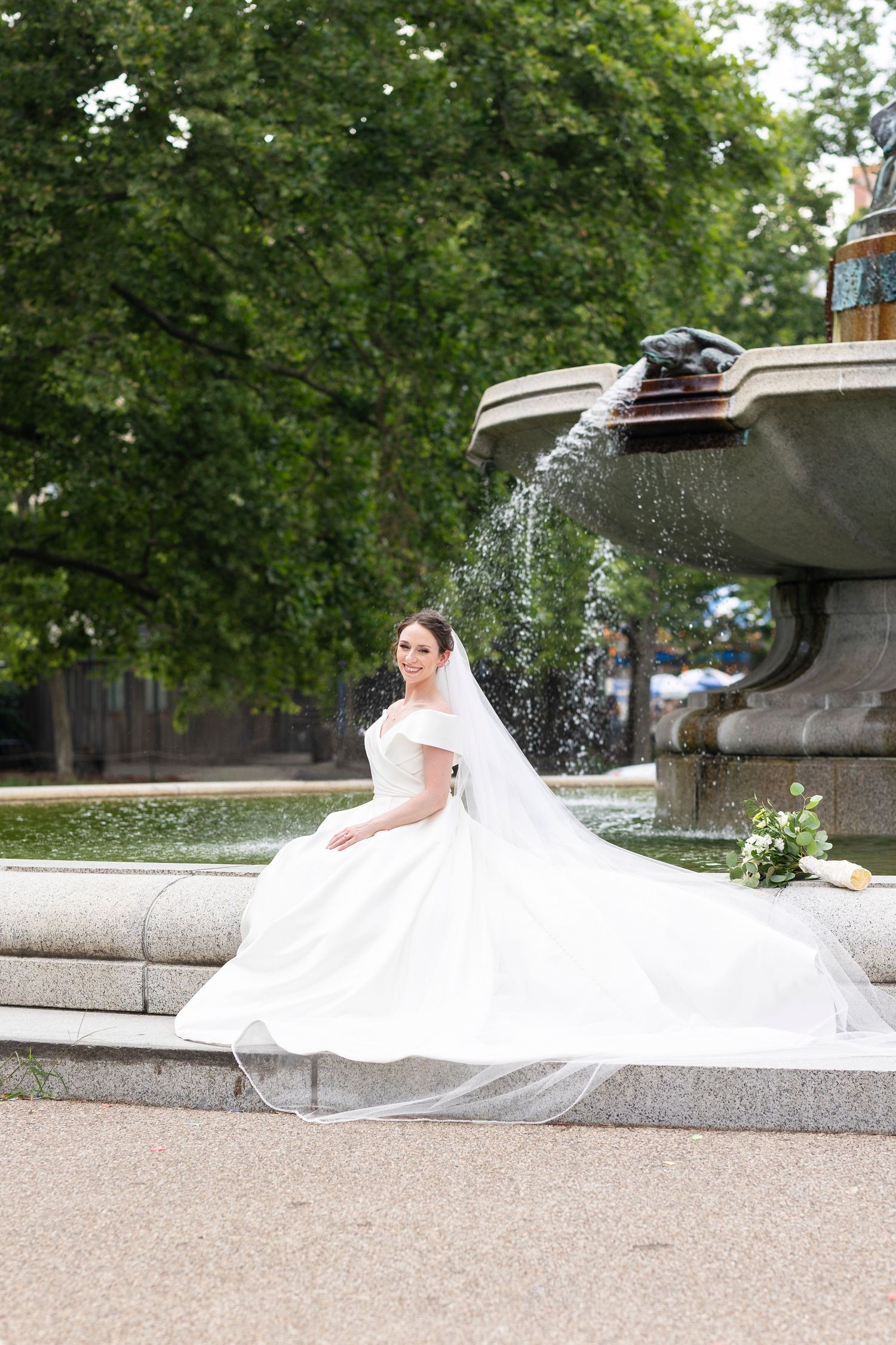 Bride in flowing white dress poses by ornate stone fountain in a garden setting.