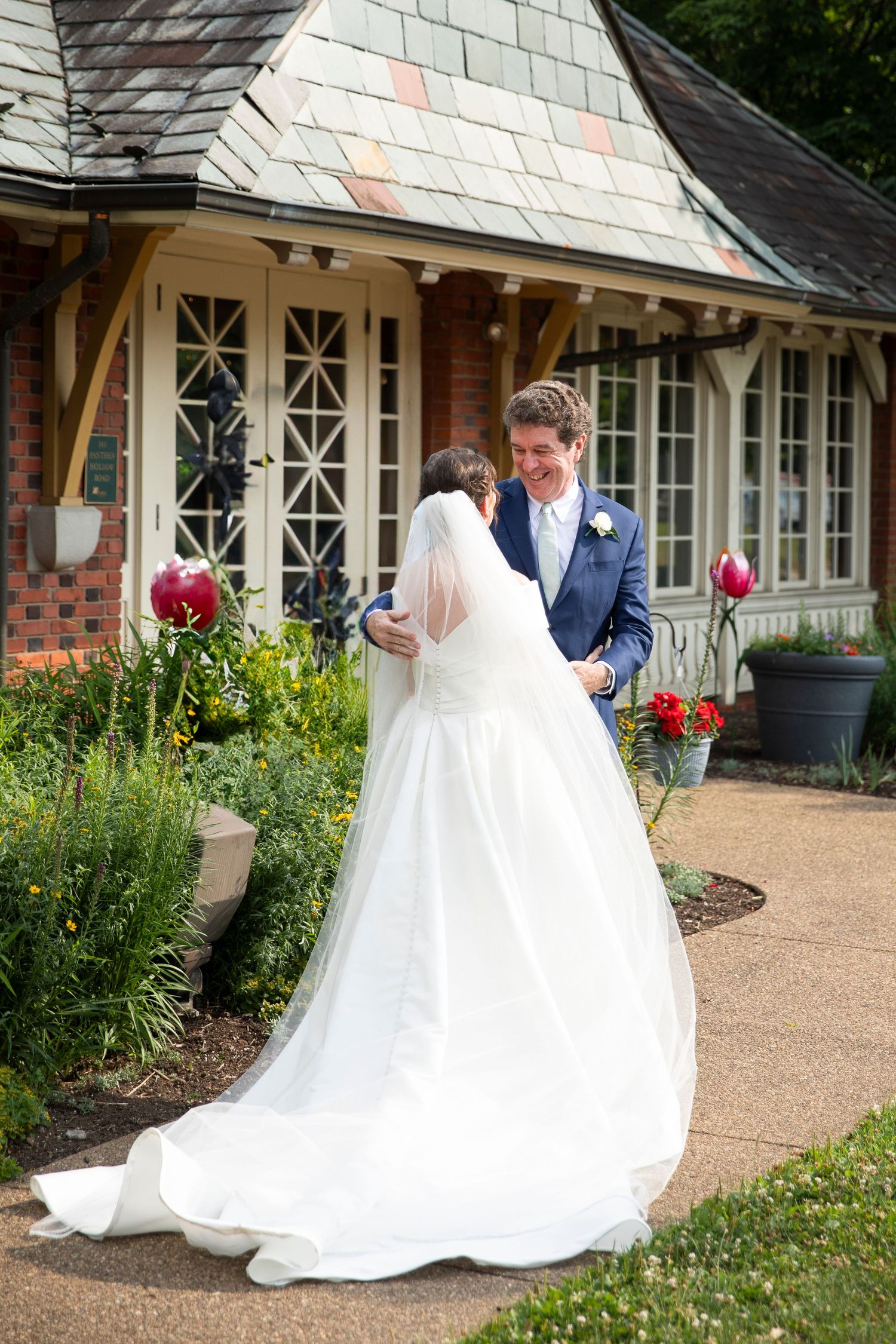 A wedding couple poses together on a paved walkway surrounded by garden flowers and brick architecture.