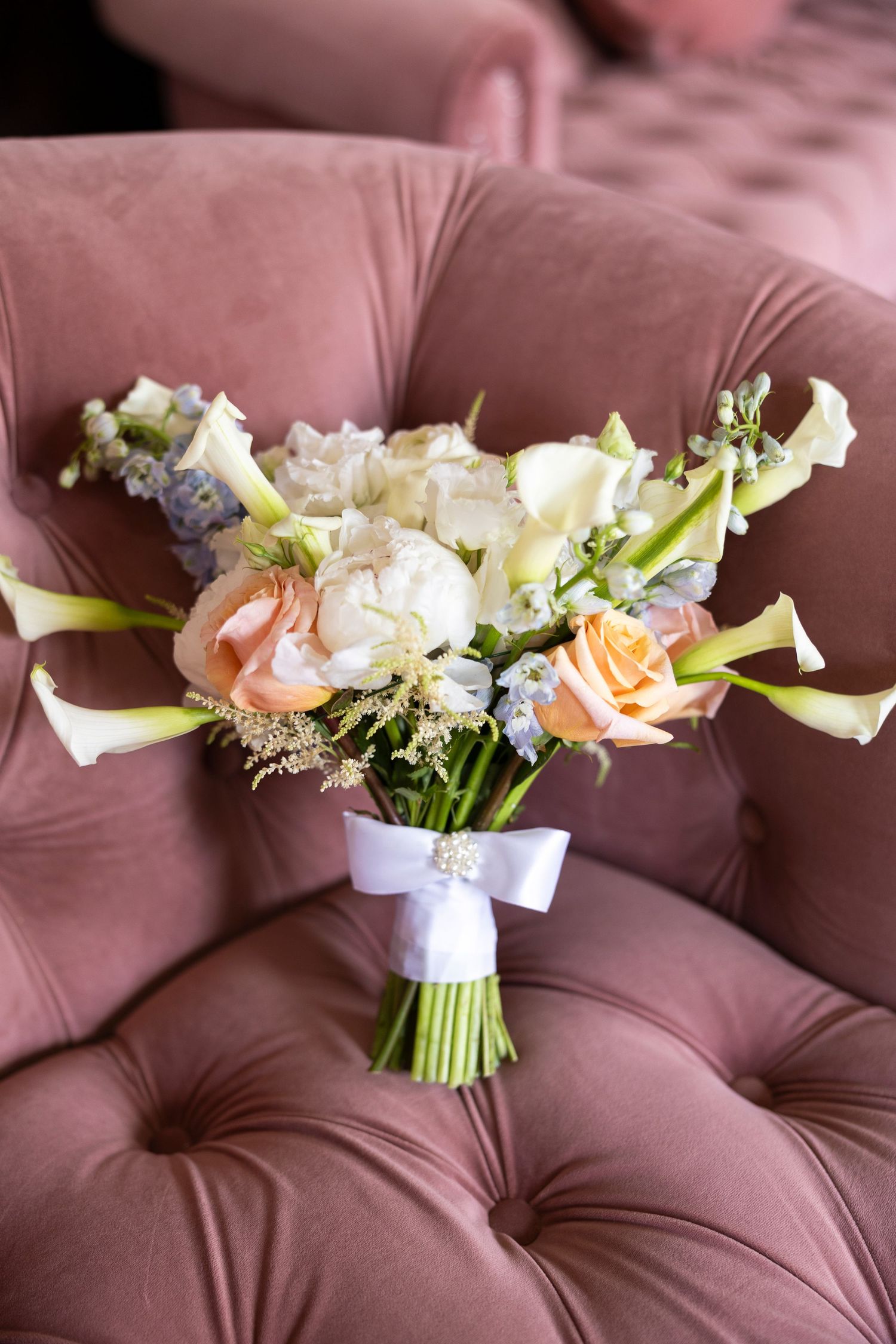 Wedding bouquets with white and peach flowers wrapped in white ribbon displayed on brown upholstered furniture.