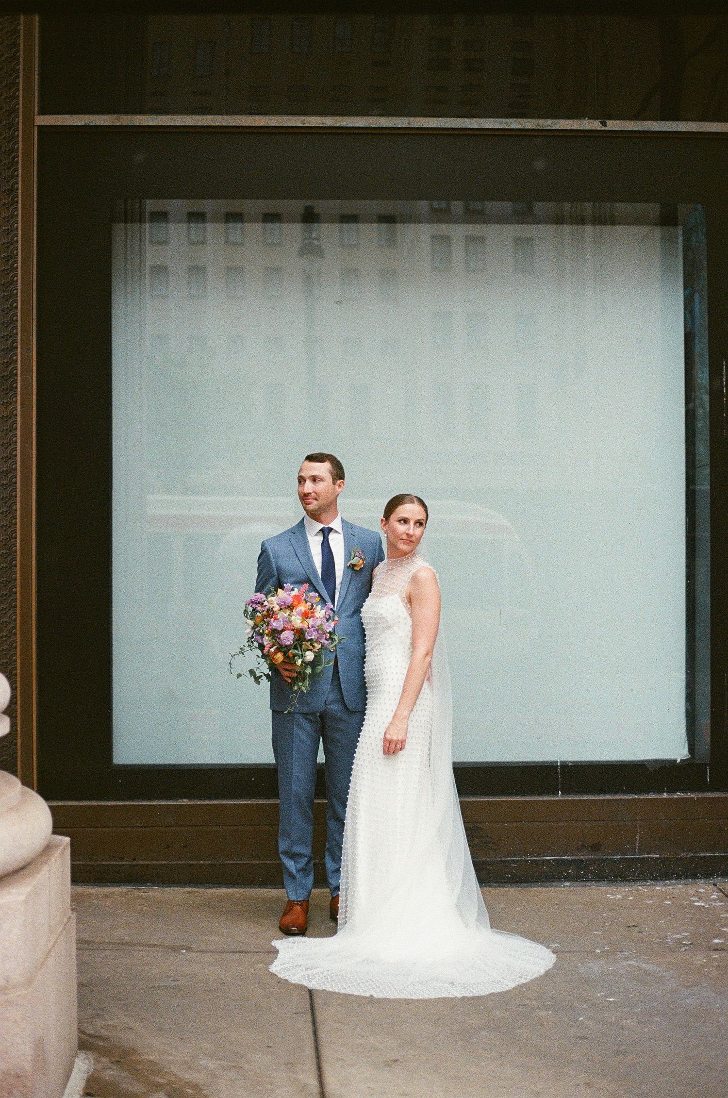 Wedding couple posing against minimalist storefront with large windows.