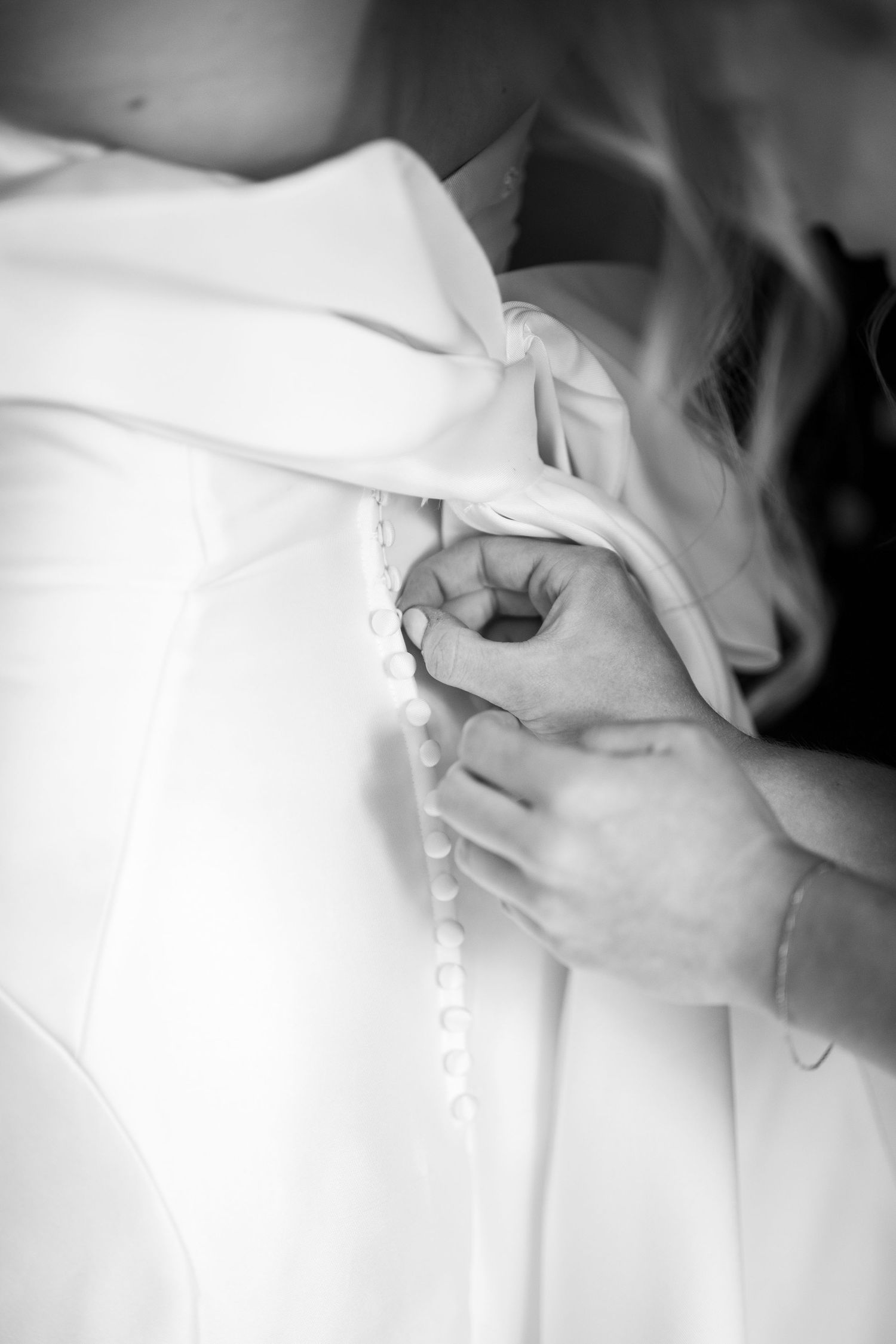 Hands buttoning the back of a white wedding dress with delicate buttons and bow detail, black and white photo.