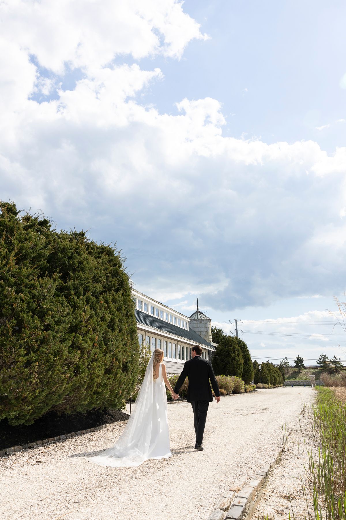Bride and groom walking hand in hand along a gravel path toward an elegant white venue surrounded by lush greenery.