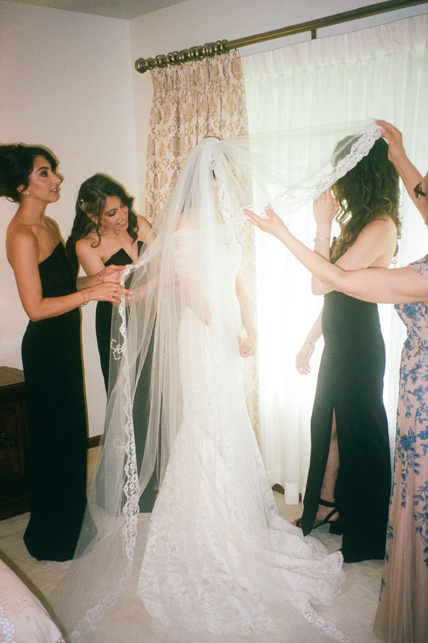 Bridesmaids in black dresses help with a wedding gown and veil near a sunlit window.