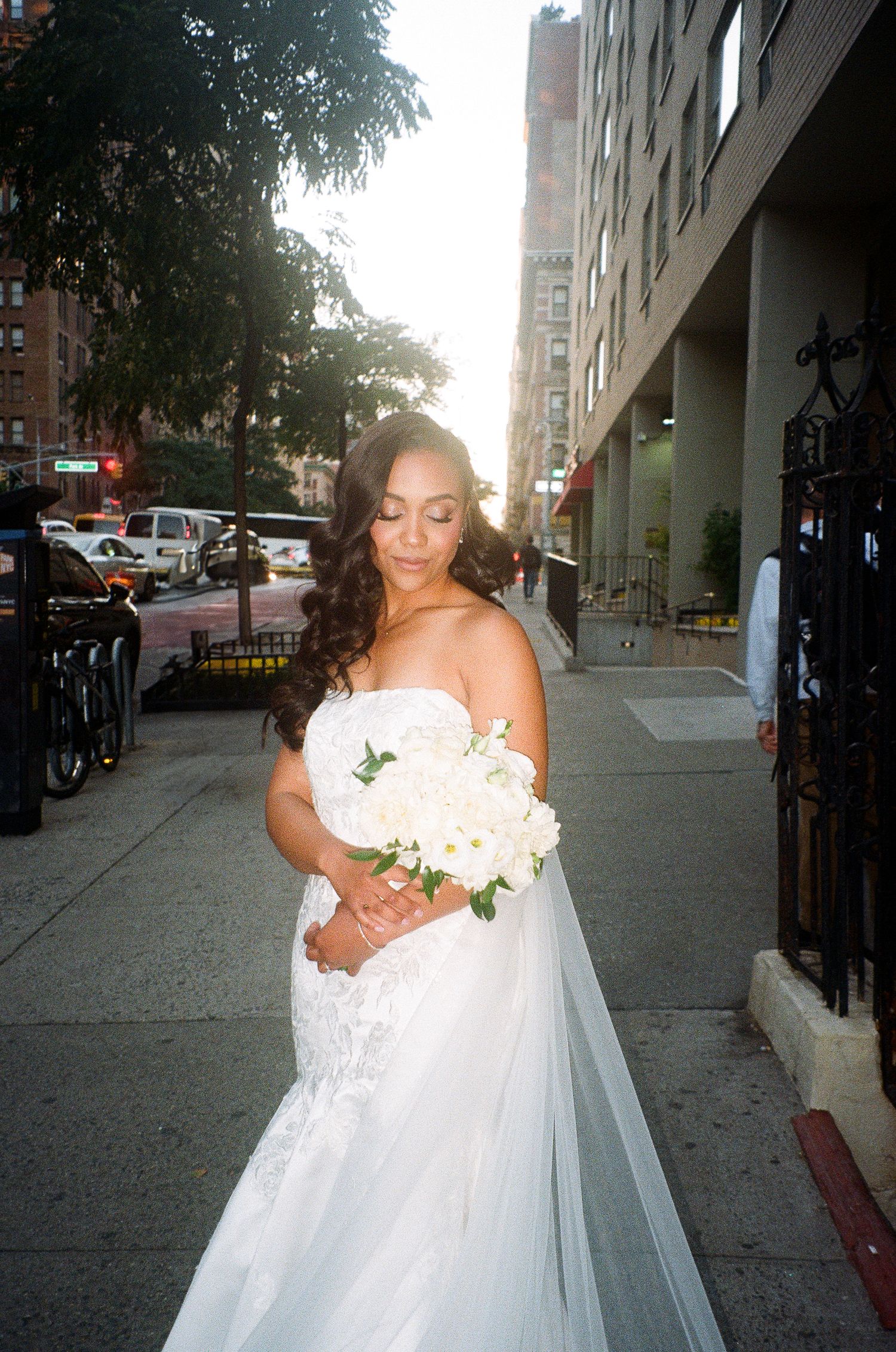 A bride in a white gown walks along an urban sidewalk holding a bouquet.