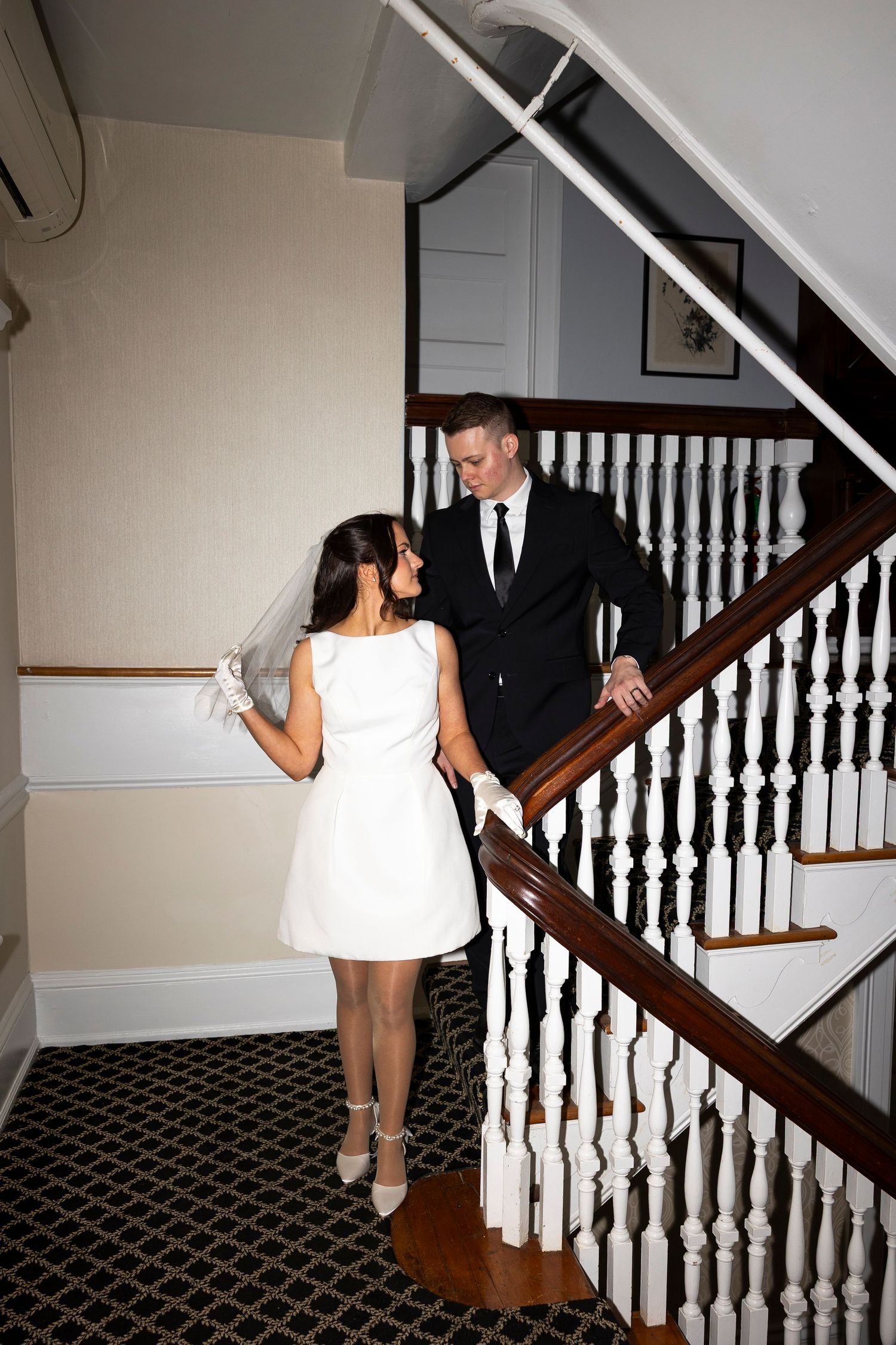 A couple stands on carpeted stairs with white bannisters in an elegant interior.