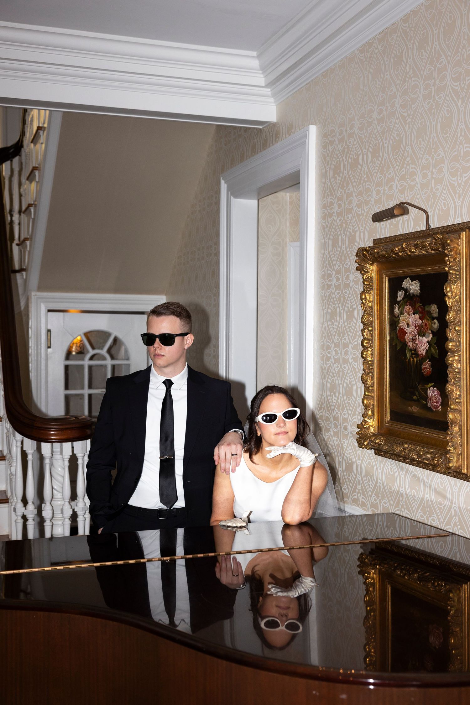 A stylish couple wearing sunglasses pose together at a piano in an elegant room.