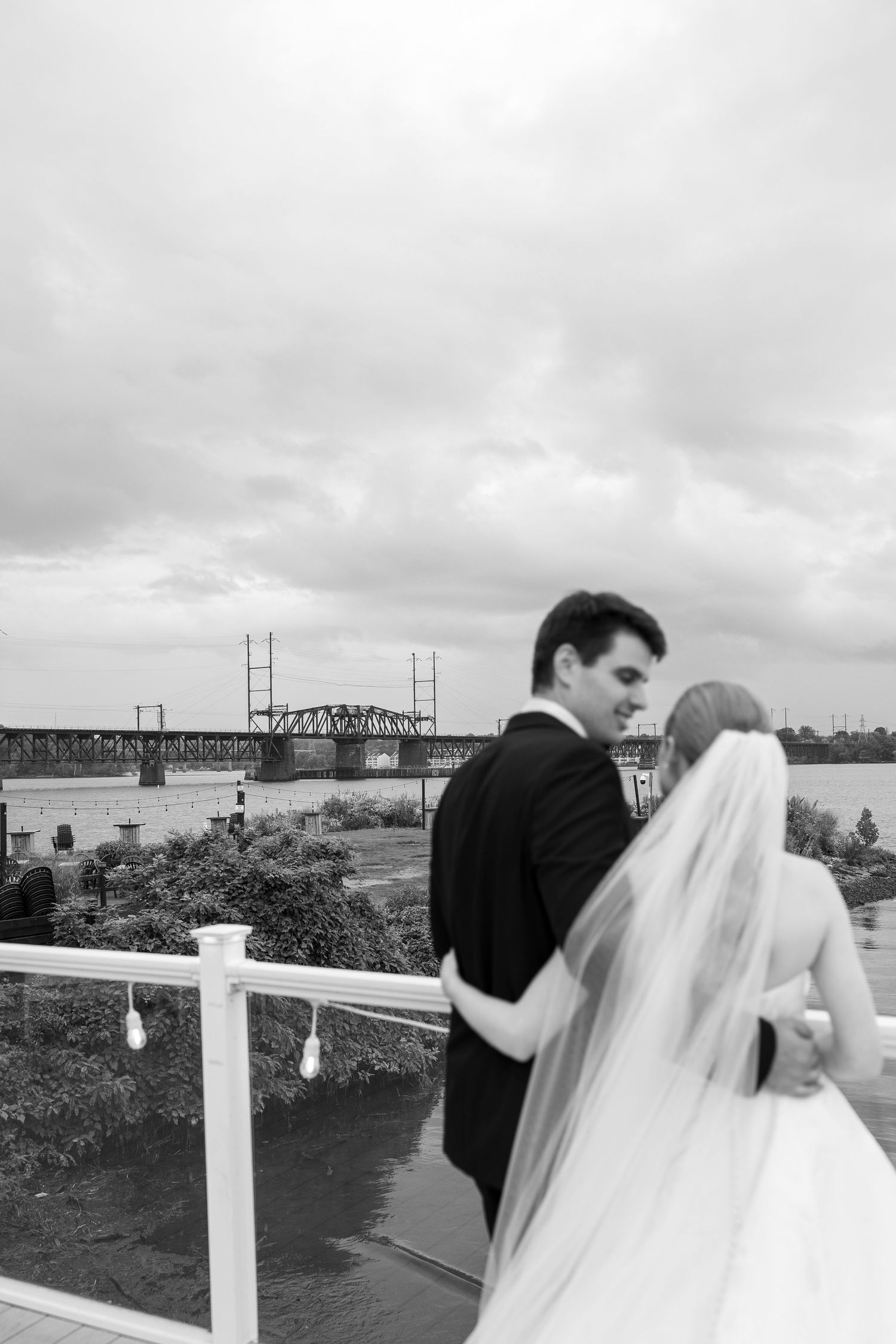 A couple in wedding attire embraces on a waterfront deck with a bridge visible in the background.