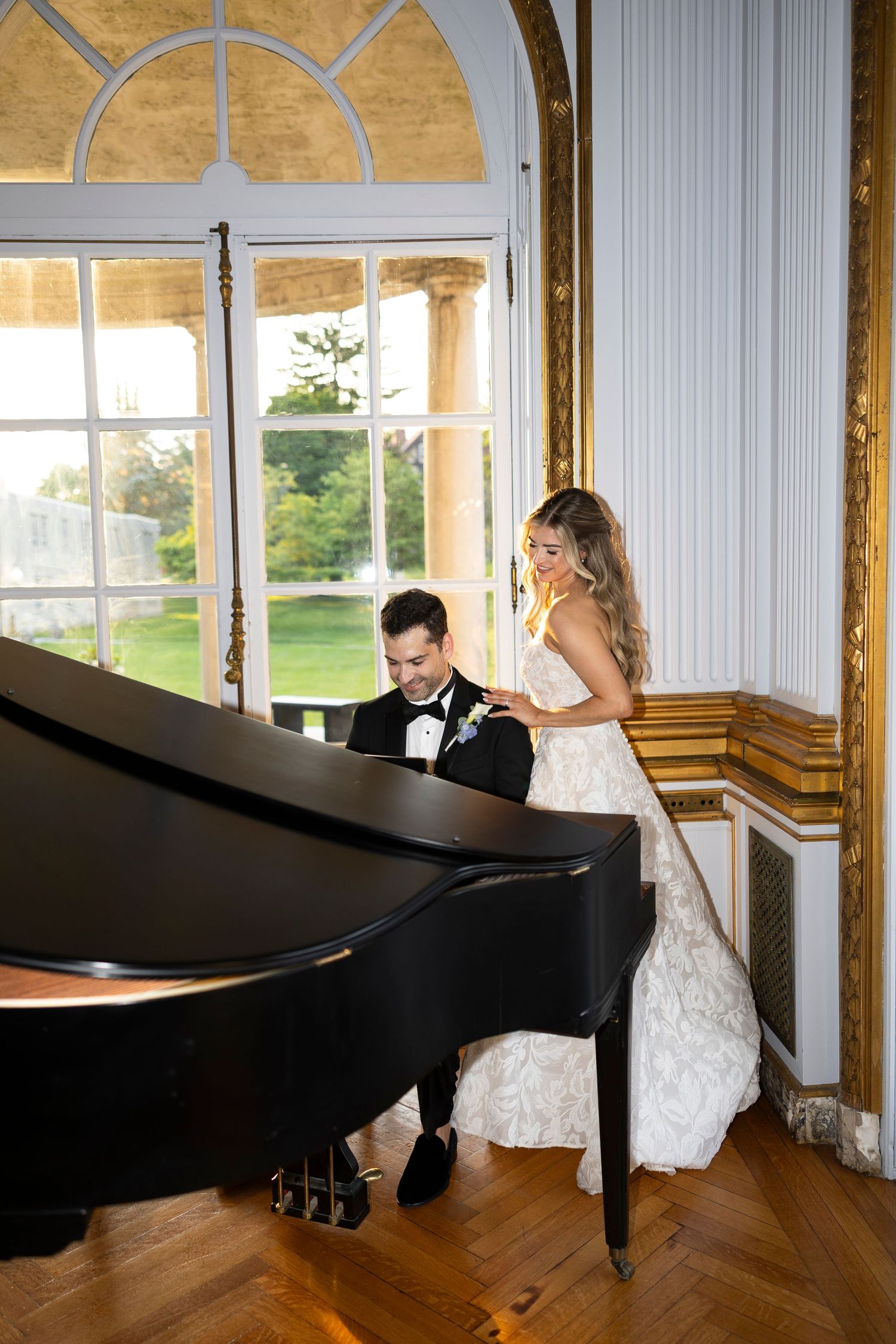 Wedding couple poses playfully with a grand piano in a sunlit room.