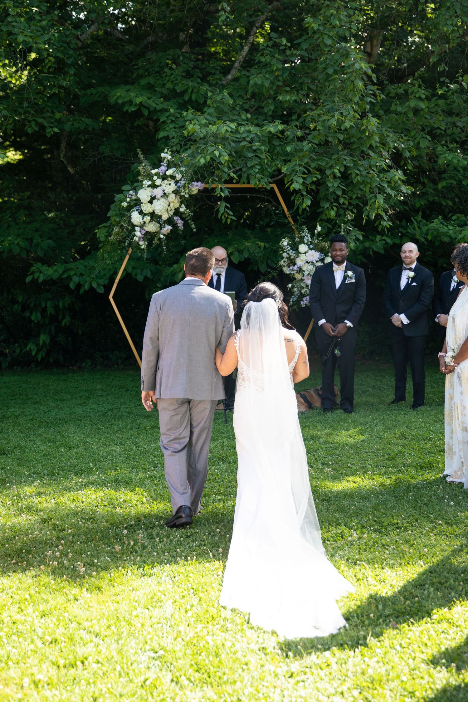 Wedding ceremony participants stand on a sunlit lawn with trees in the background.