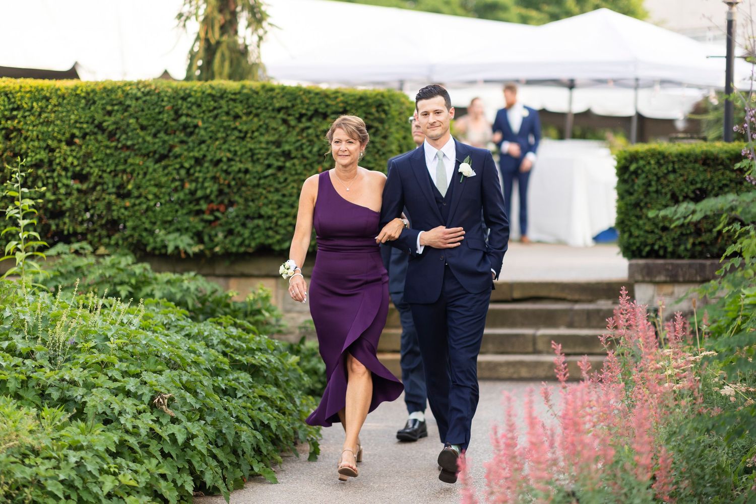 Wedding party members in purple and navy attire walk along a garden path.