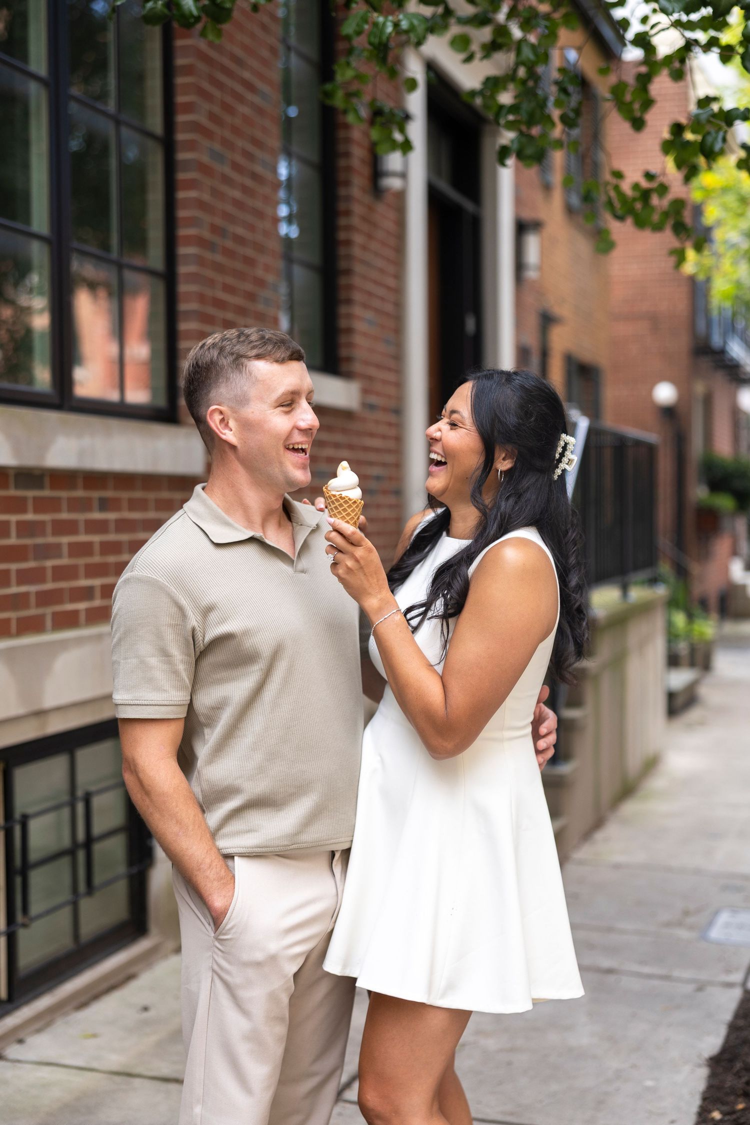 A couple shares joyful laughter while enjoying ice cream cones on a city sidewalk.