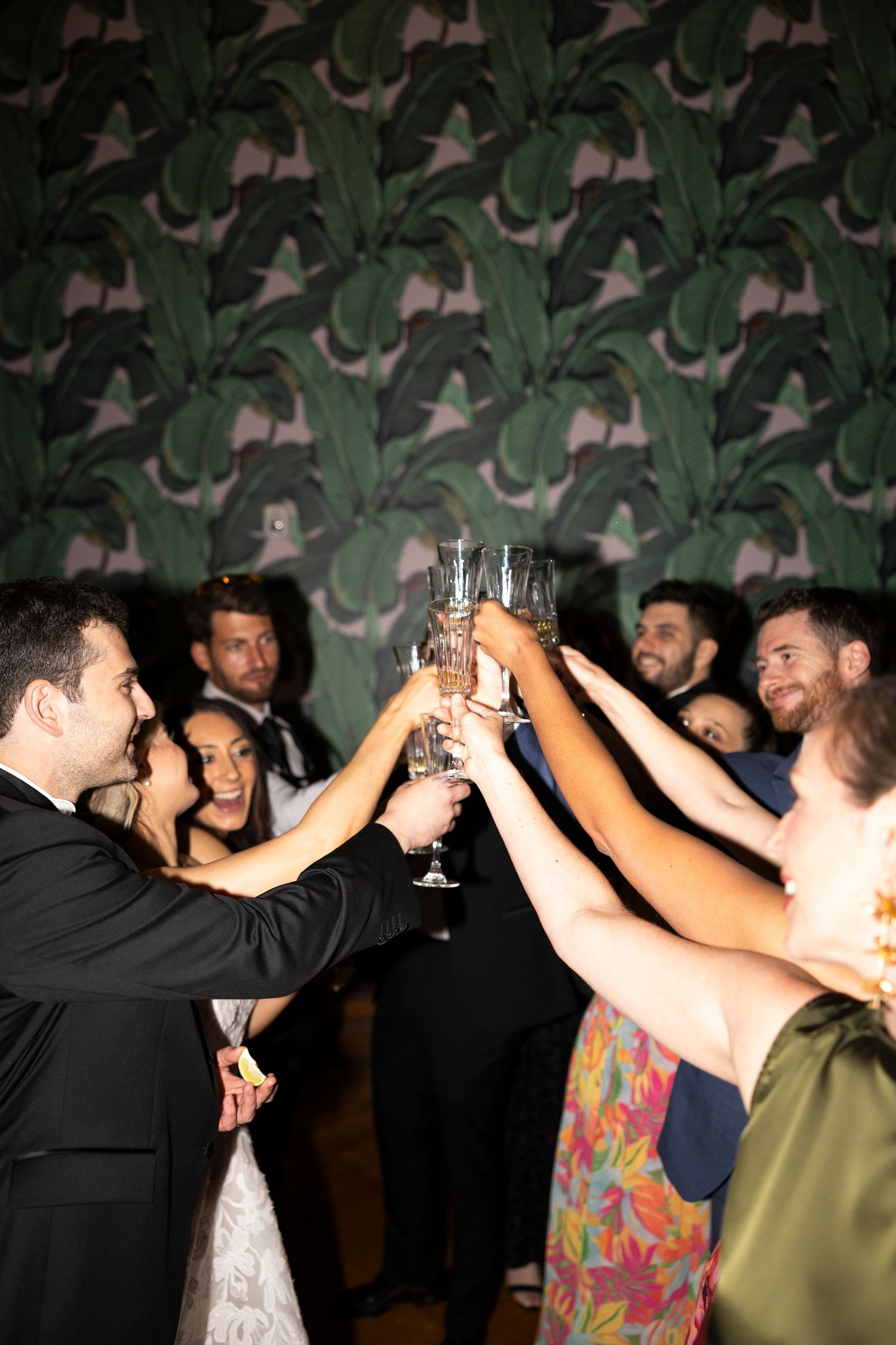 Wedding guests raising champagne glasses in a celebratory toast at a reception with tropical leaf wallpaper backdrop.